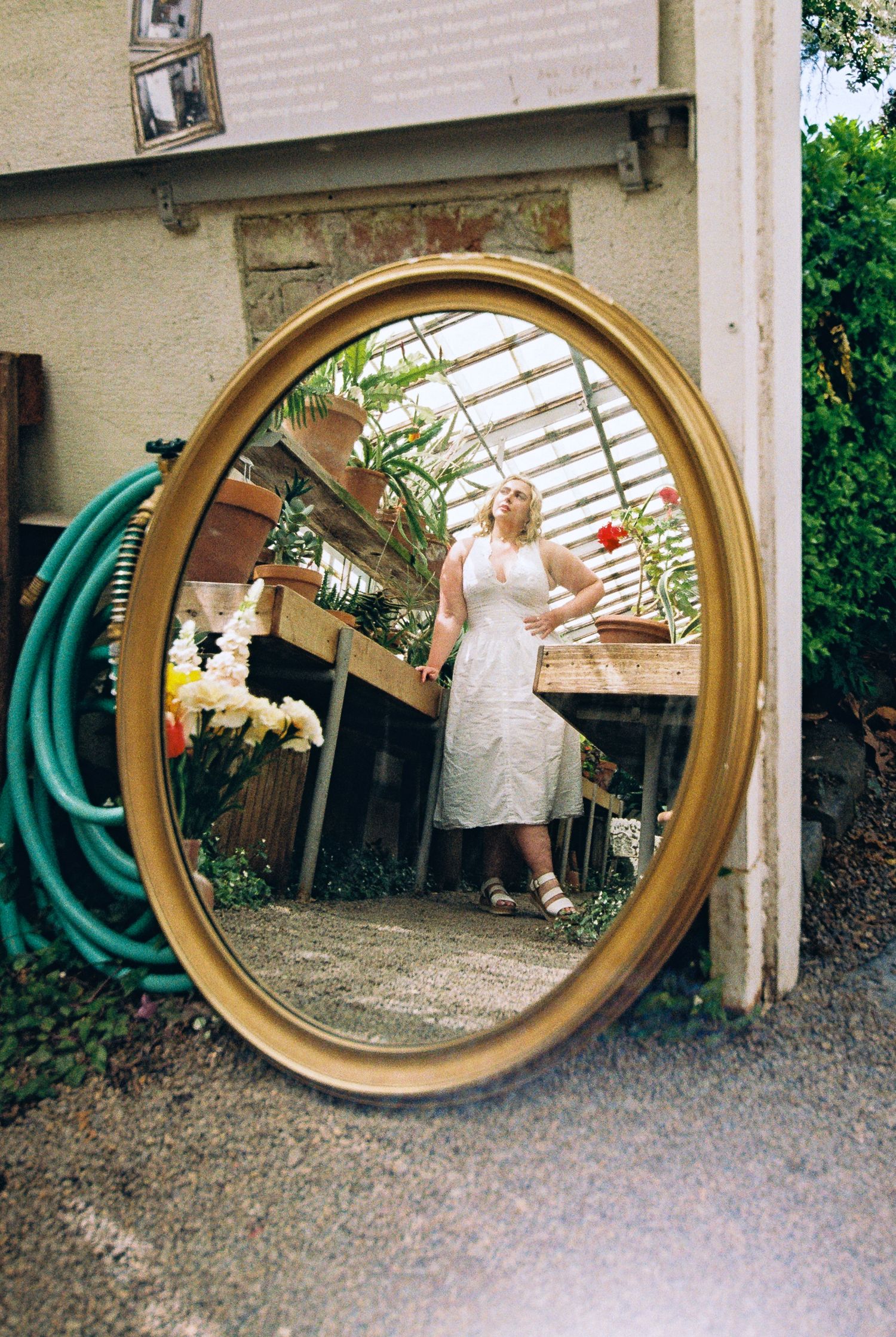 Woman in white dress reflected in ornate gold oval mirror outdoors near greenhouse.