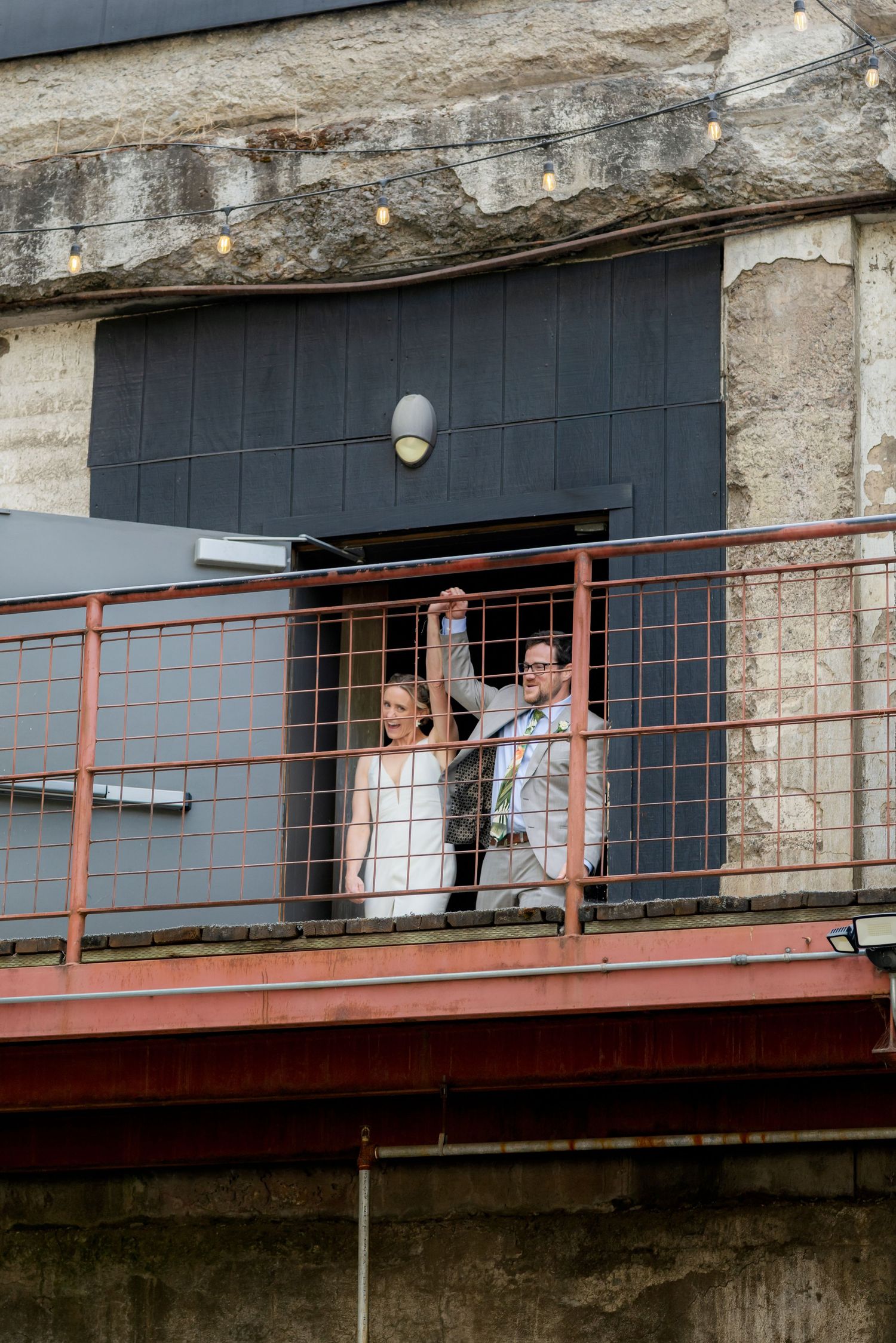 Couple celebrating on a rustic industrial balcony with string lights overhead.