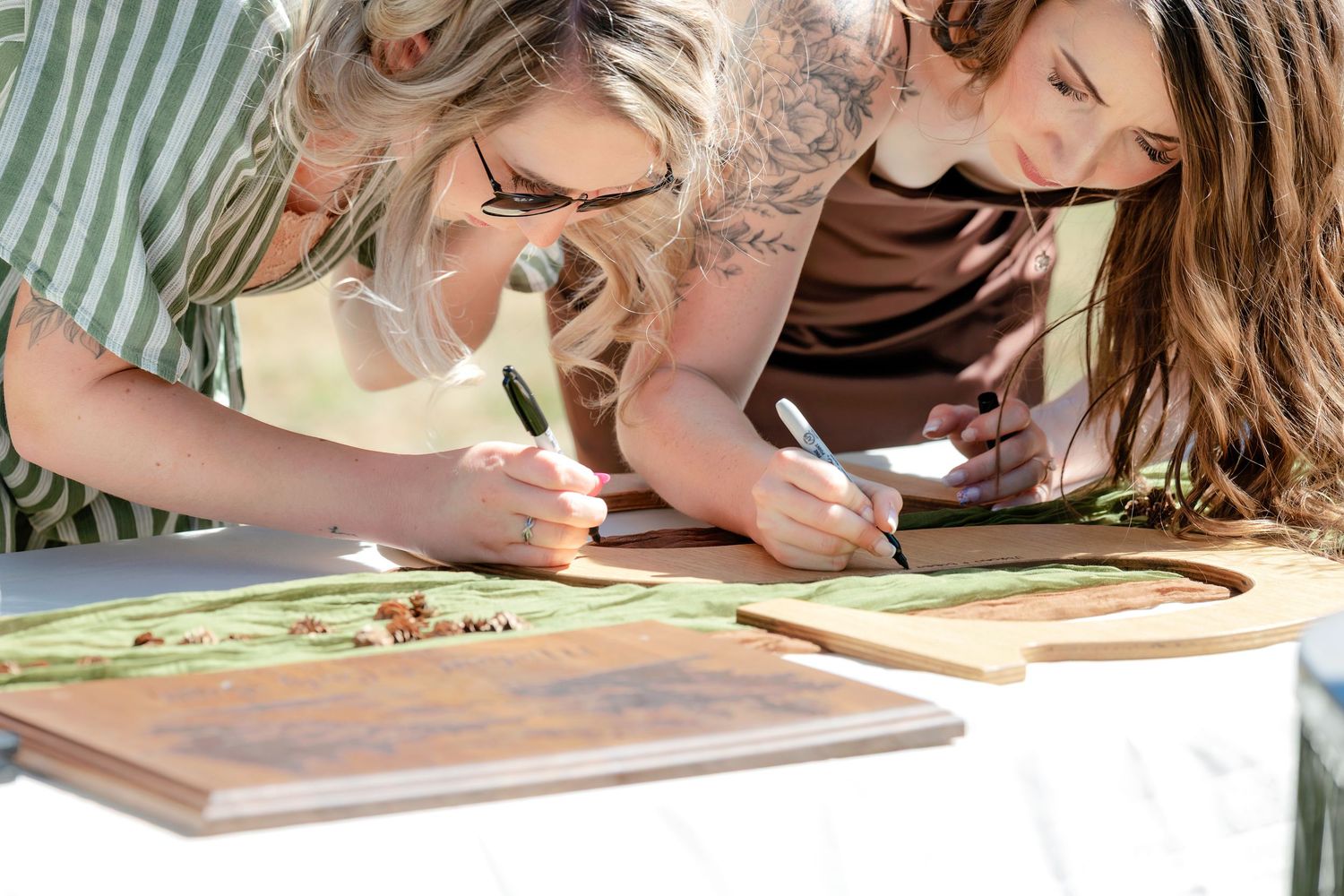 Three women leaning over a table signing wooden boards at an outdoor wedding event.