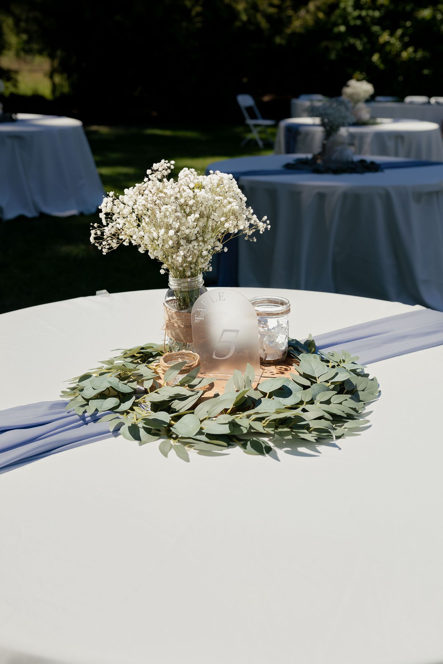 Elegant outdoor wedding reception table with baby's breath centerpiece, eucalyptus wreath, and lavender ribbon runner.