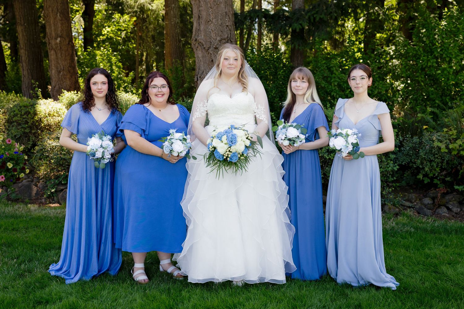Bride in white gown with four bridesmaids in blue dresses holding bouquets outdoors.