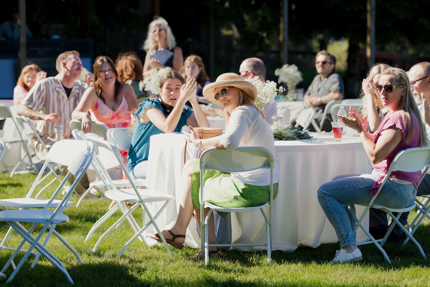 Wedding guests seated at white tables on grass enjoying outdoor daytime reception celebration.
