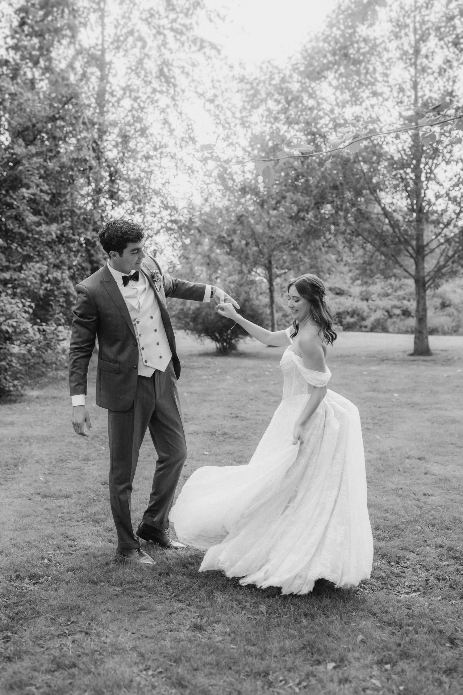 Groom twirls bride in flowing white gown on grassy lawn in romantic black and white wedding portrait.