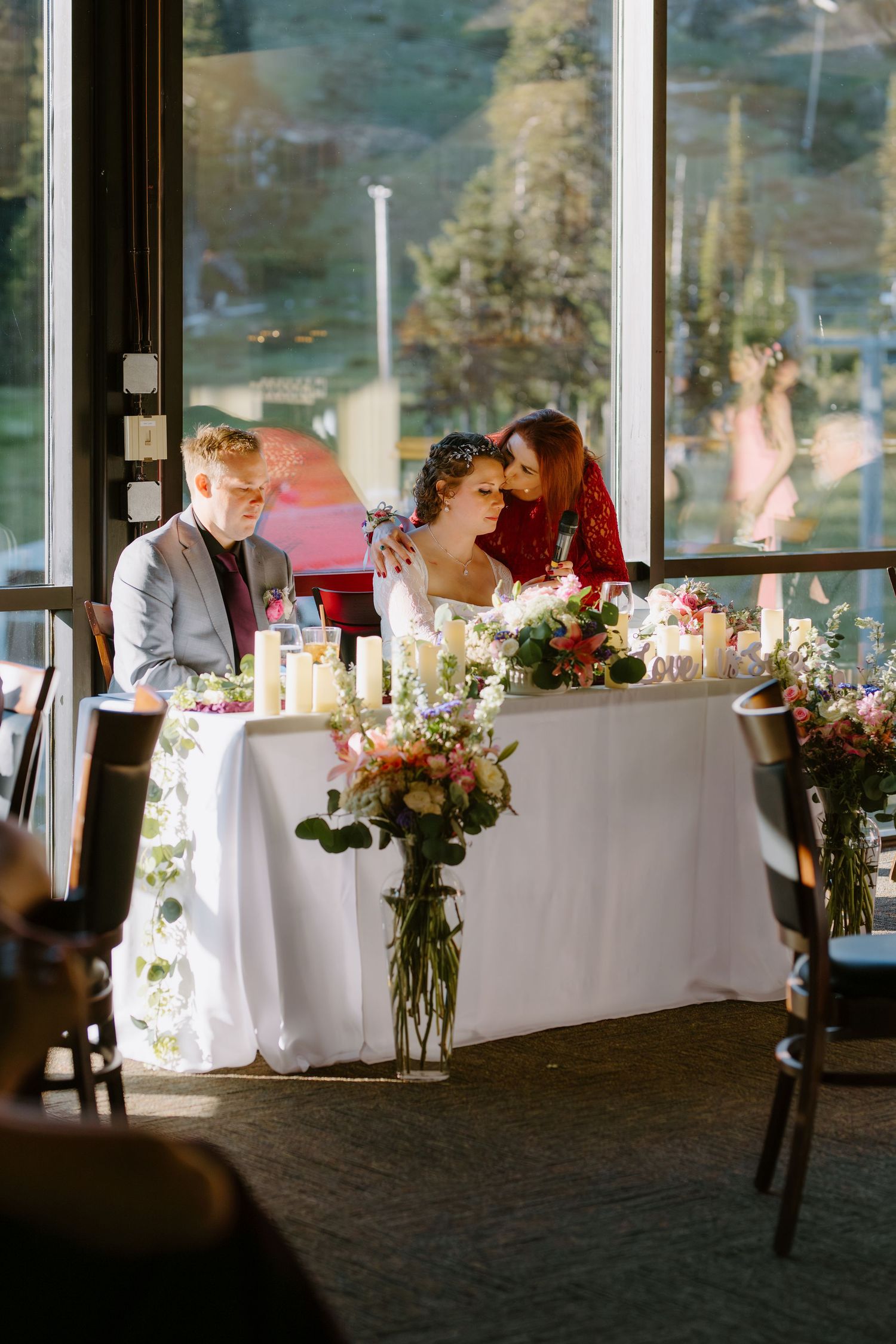 Bride and groom seated at floral-decorated sweetheart table with large mountain view windows behind them.