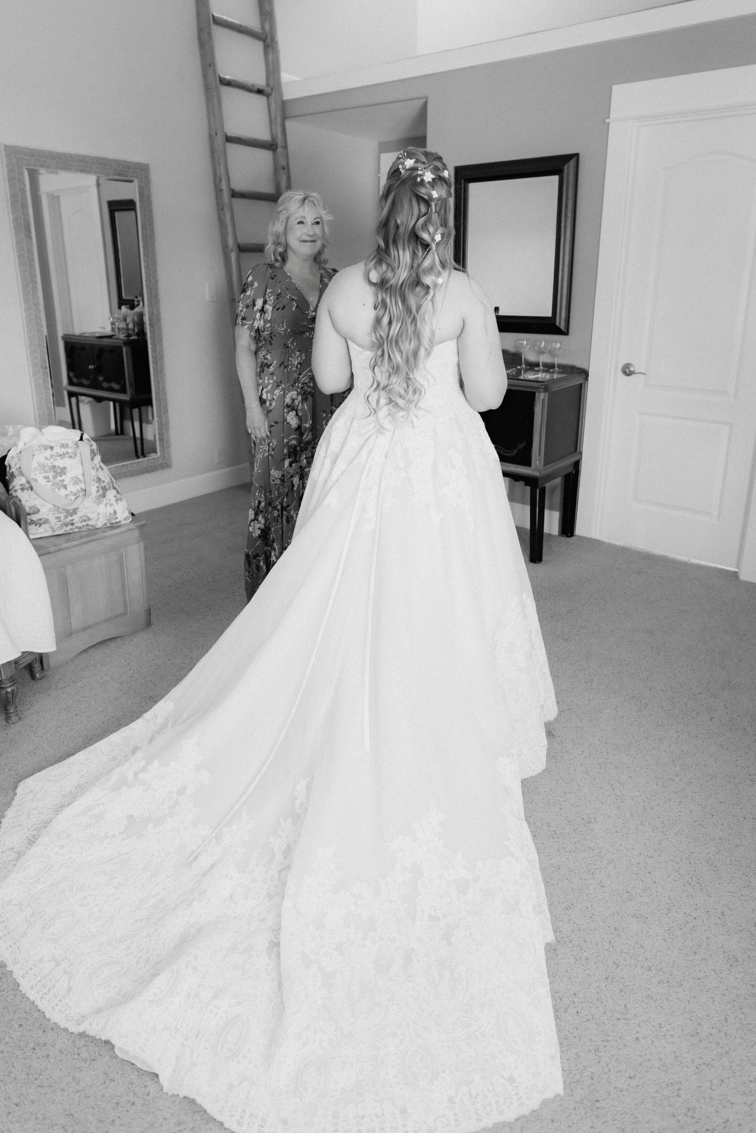 Black and white photo of a bride in a long lace wedding gown with a train, viewed from behind in a bridal suite.