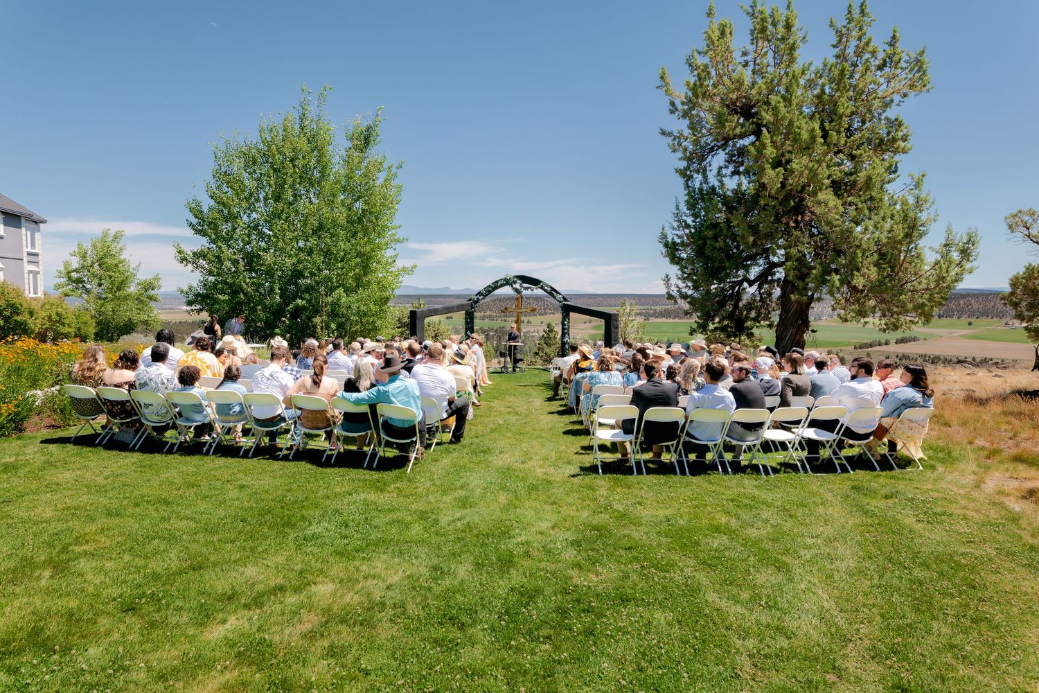 Outdoor wedding ceremony with guests seated on white chairs on a green lawn under a sunny blue sky.