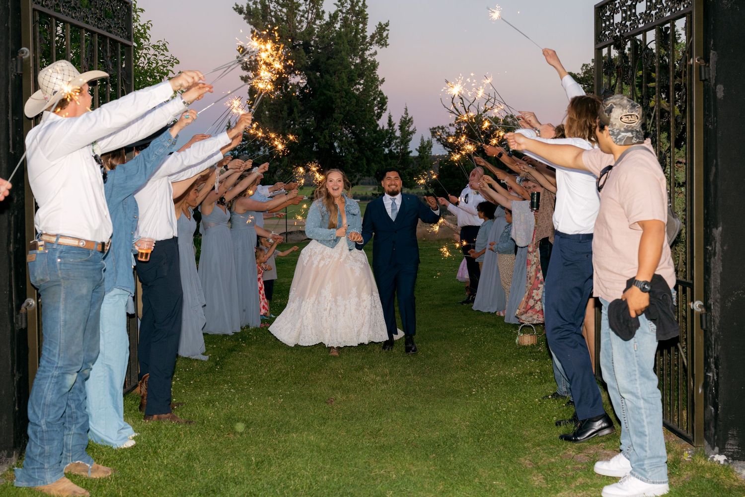 Bride and groom exit through sparkler tunnel surrounded by cheering wedding guests at dusk.