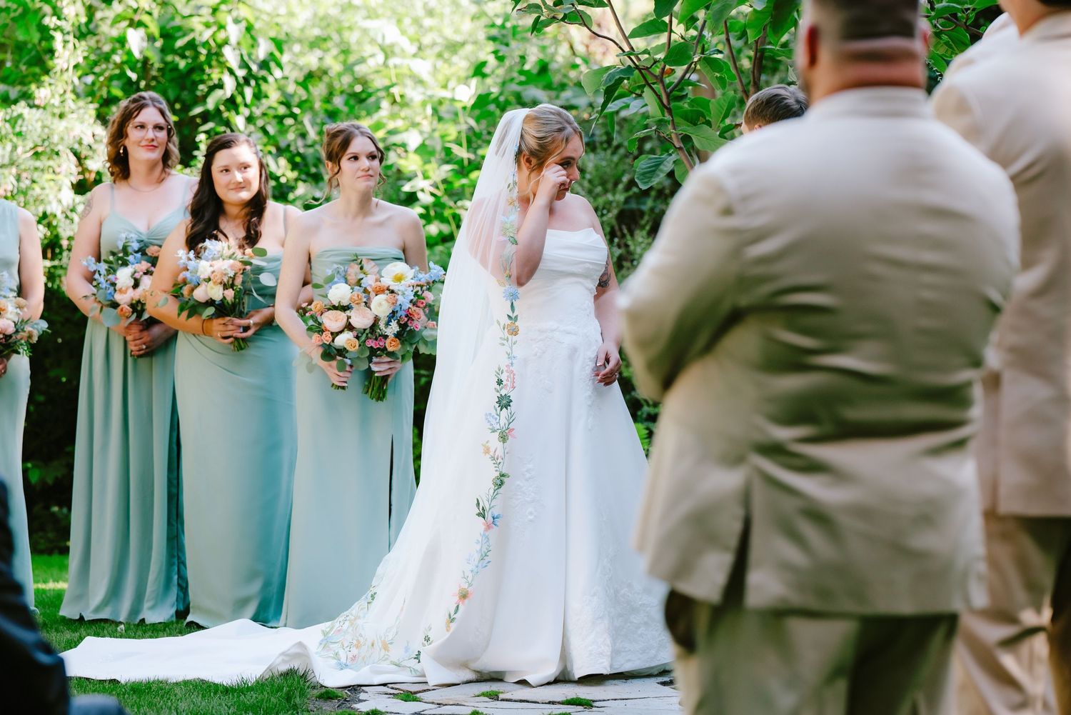 Bride in white gown with veil stands emotional at outdoor wedding ceremony with bridesmaids in sage green.