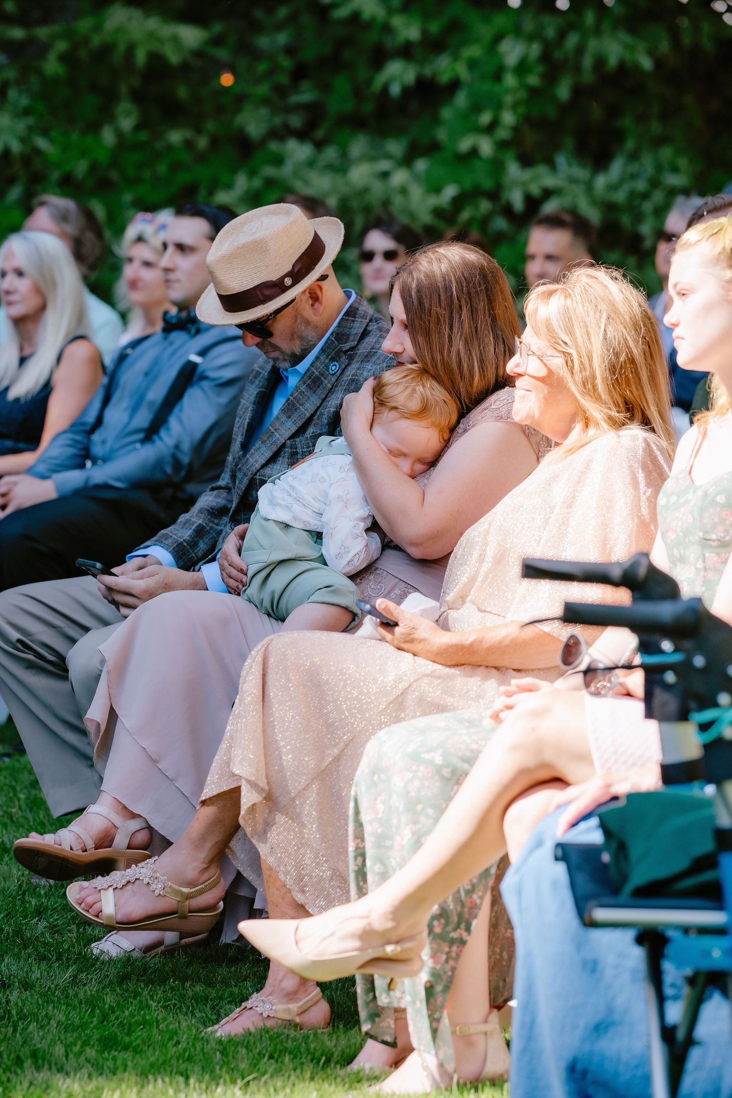 Wedding guests seated outdoors watching ceremony, woman holding baby in pink dress at garden wedding.
