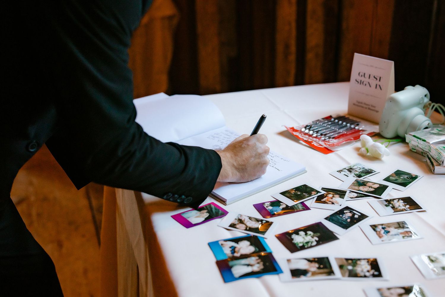 Guest signing wedding guest book surrounded by Polaroid photos on decorated table.