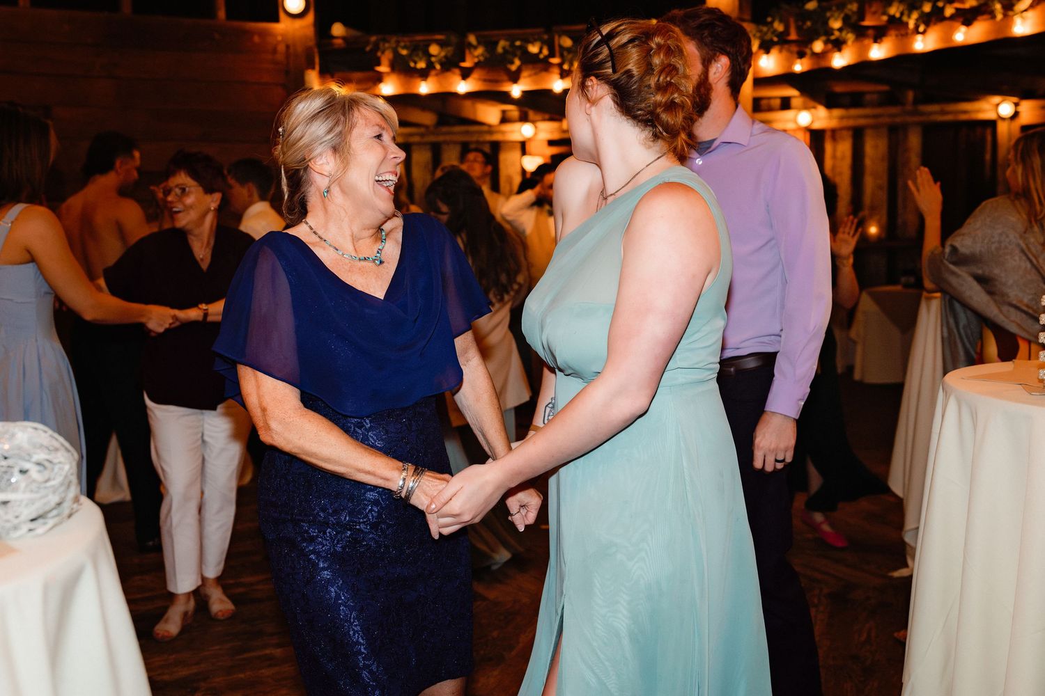 Two women in elegant dresses hold hands and dance together at a warmly lit wedding reception.