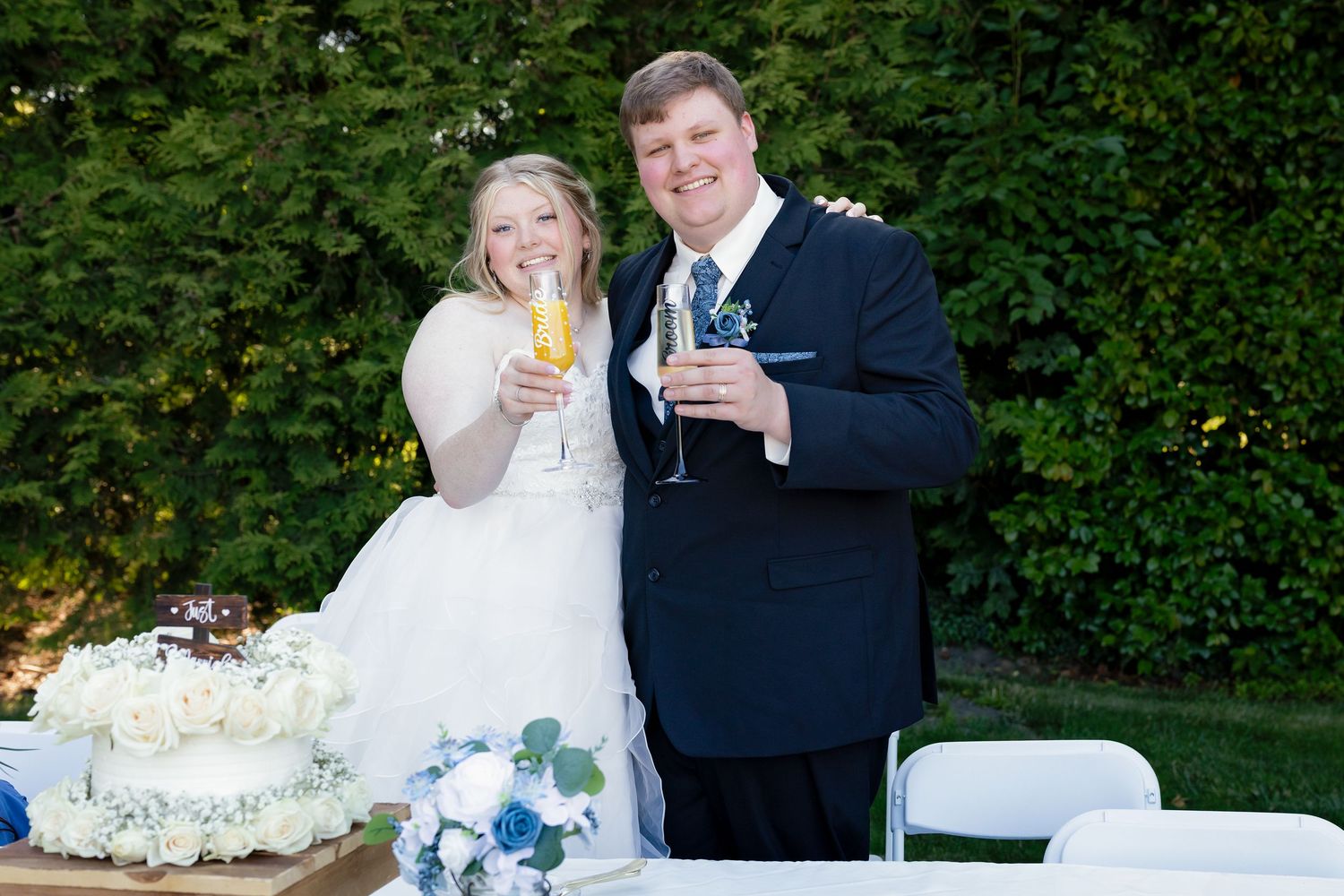 Newlywed couple toasting with champagne glasses at outdoor wedding reception with white cake and floral arrangements.