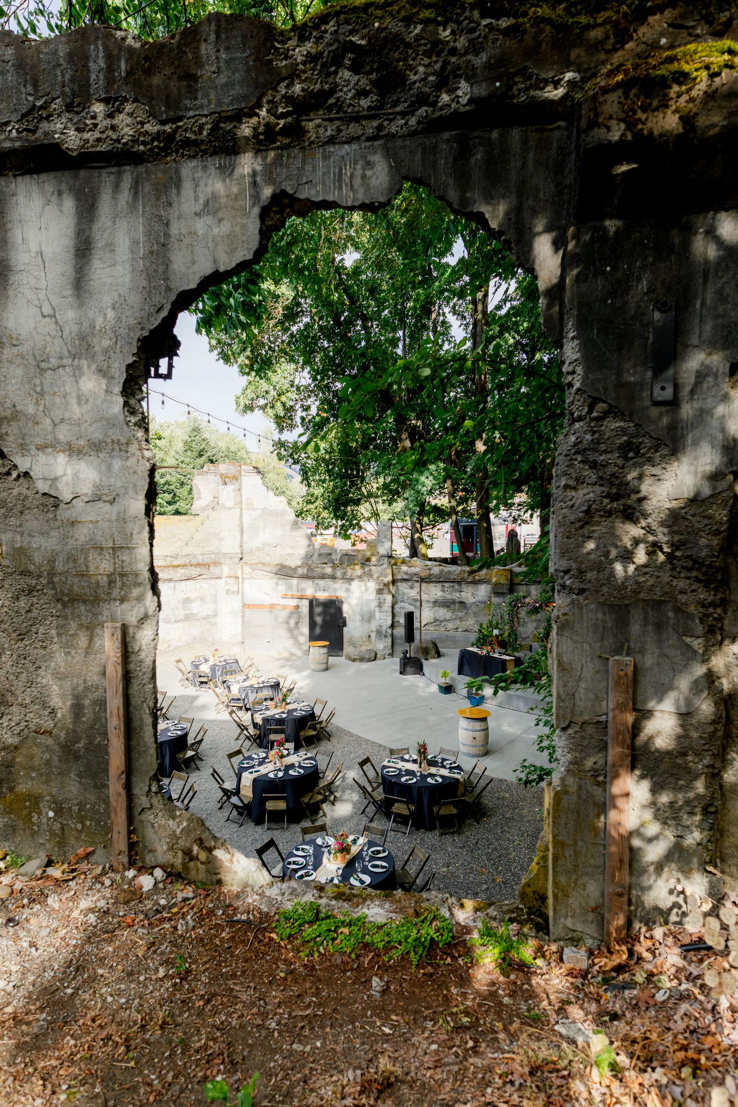 Rustic outdoor event space viewed through a broken concrete wall, with elegantly set tables among lush trees.