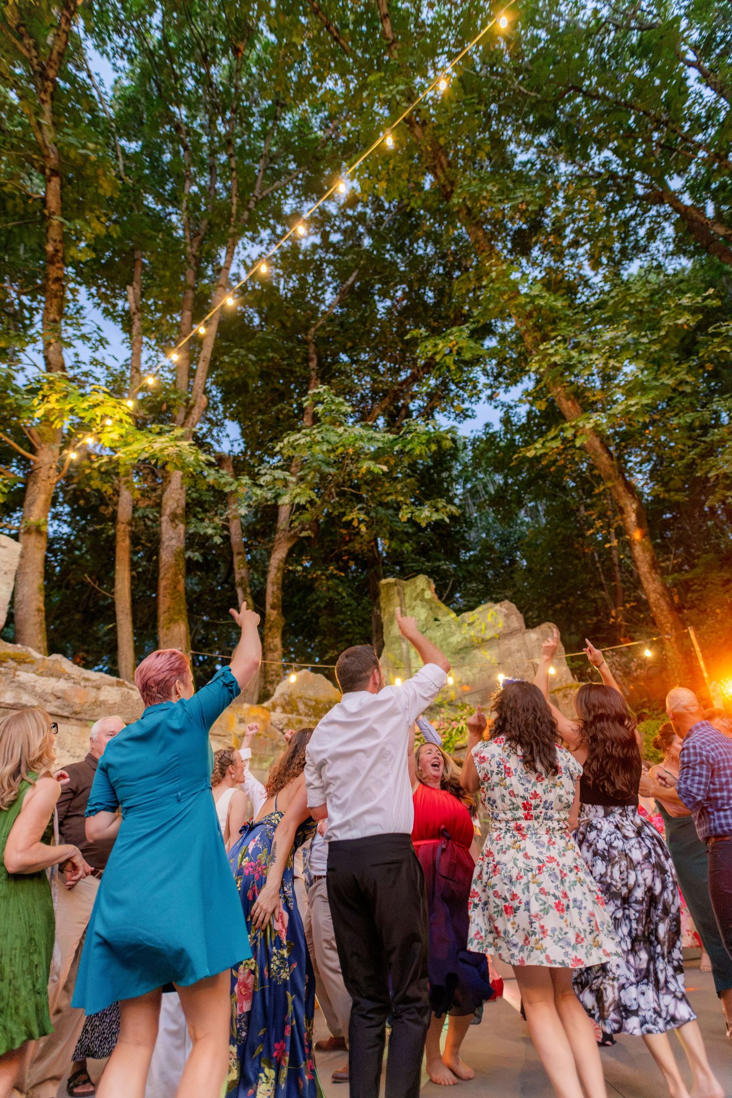 Guests dancing outdoors at a wedding reception under string lights in a lush green forest setting.