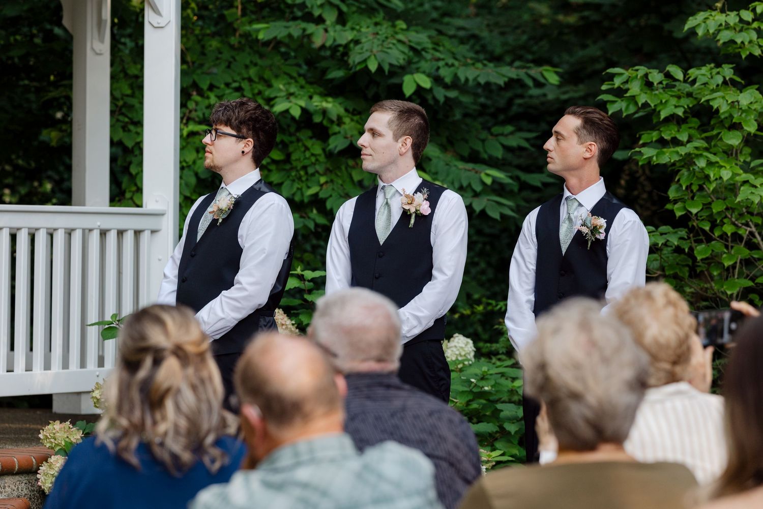 Three groomsmen in black vests and white shirts stand at outdoor wedding ceremony gazebo before seated guests.