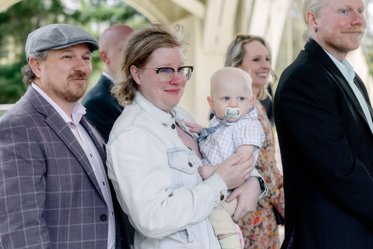 Wedding guests smile and hold a baby with a pacifier during an outdoor ceremony celebration.