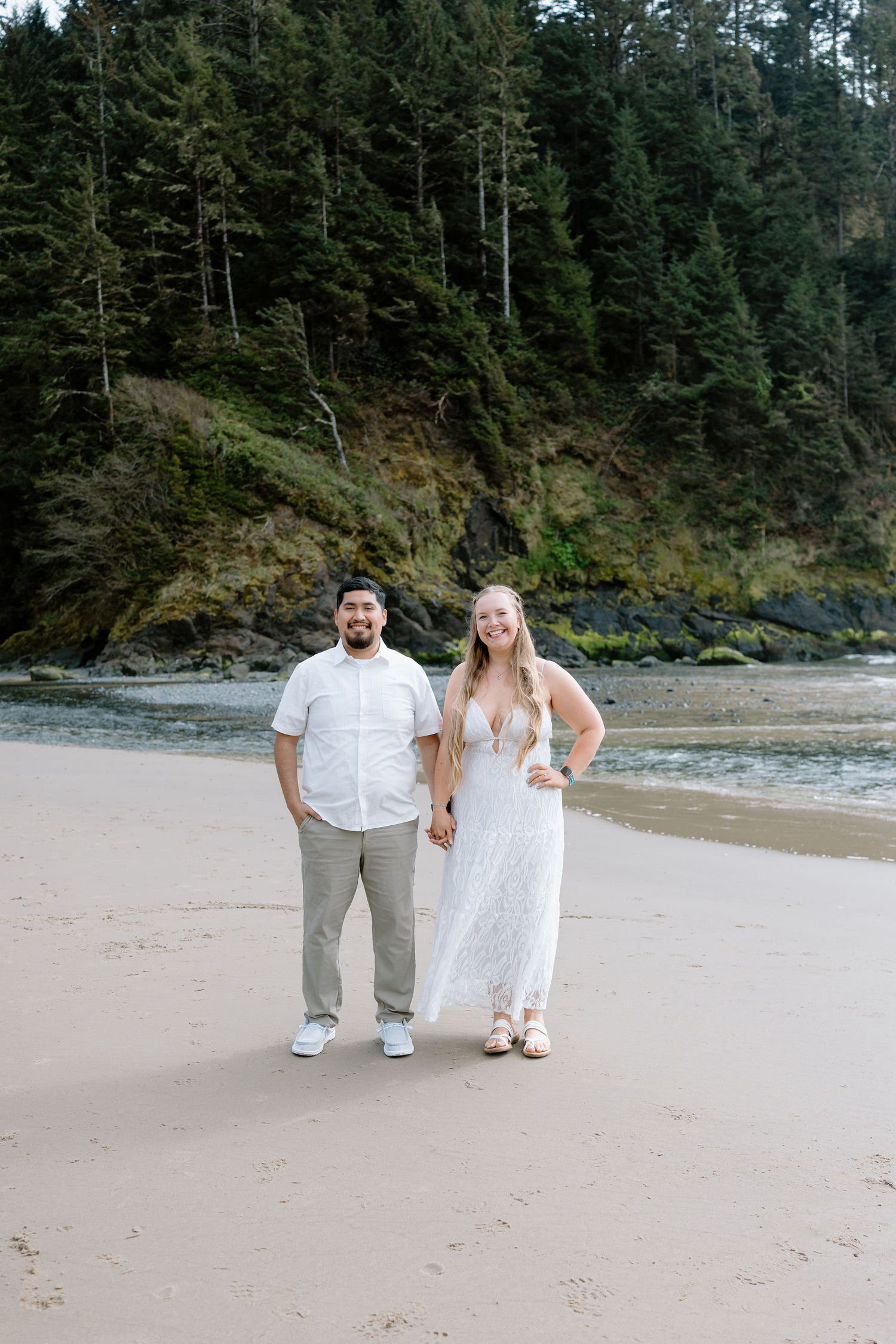 Couple in white outfits holding hands on a sandy beach with lush green forest and rocky cliffs behind them.