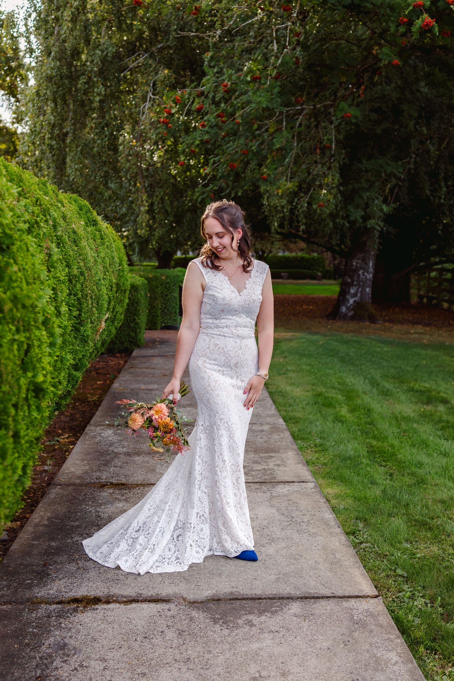 Person in white lace wedding dress adjusts their gown while standing on a stone pathway in a garden setting.