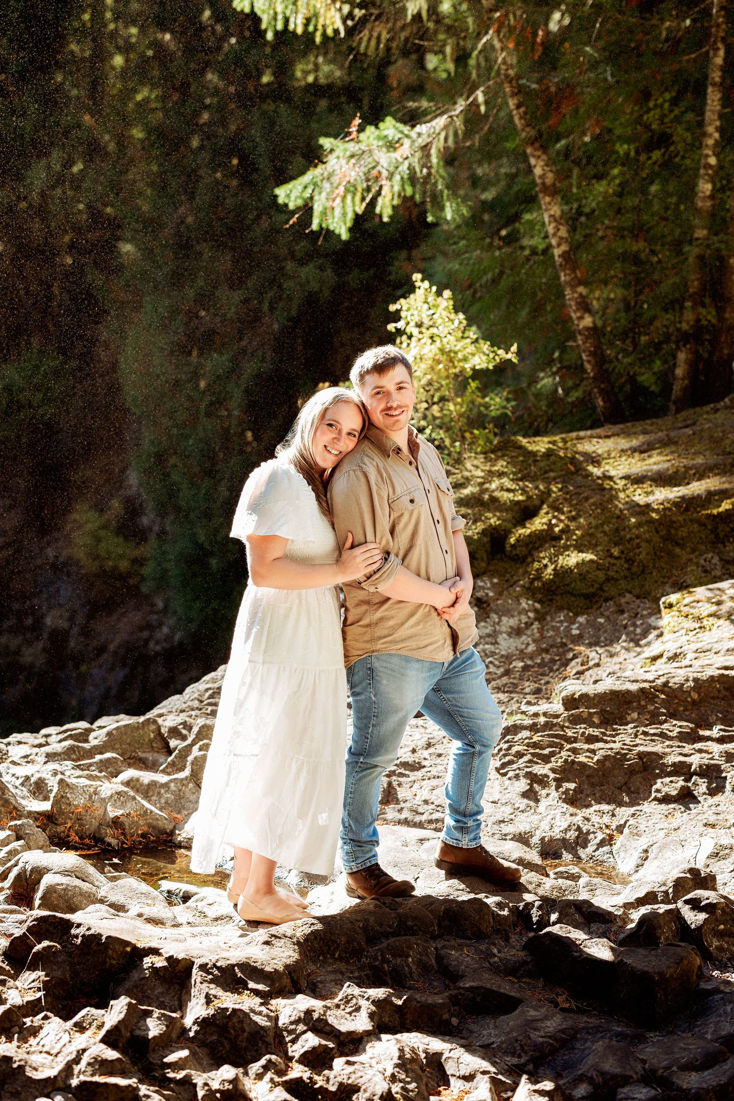A couple sits together on rocky terrain in a forest setting with dappled sunlight filtering through the trees.