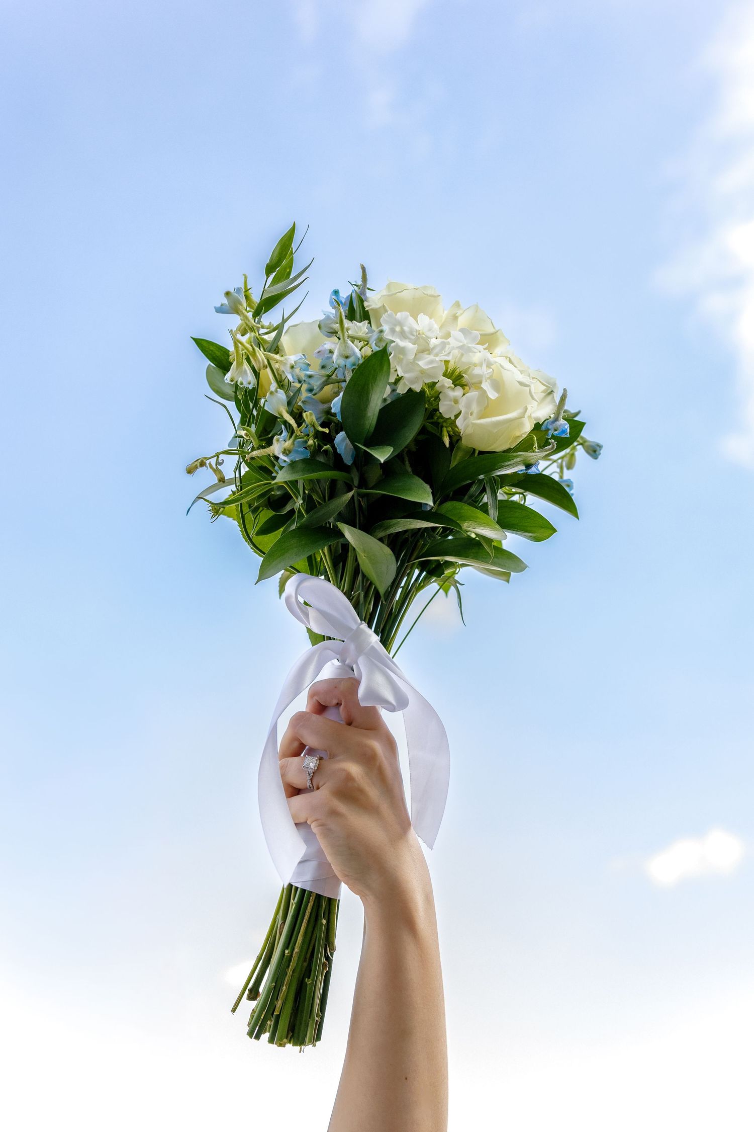 White rose and greenery wedding bouquet held up against a bright blue sky.