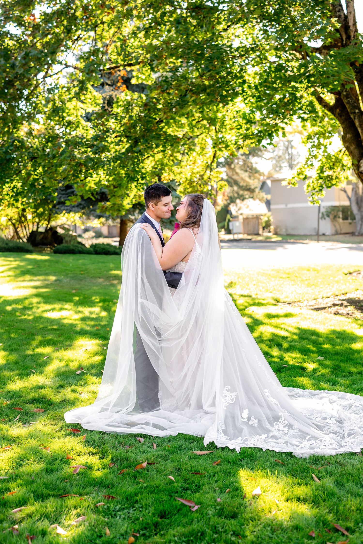 Elegant wedding couple poses on lush green grass with flowing white veil in sunlit garden setting.