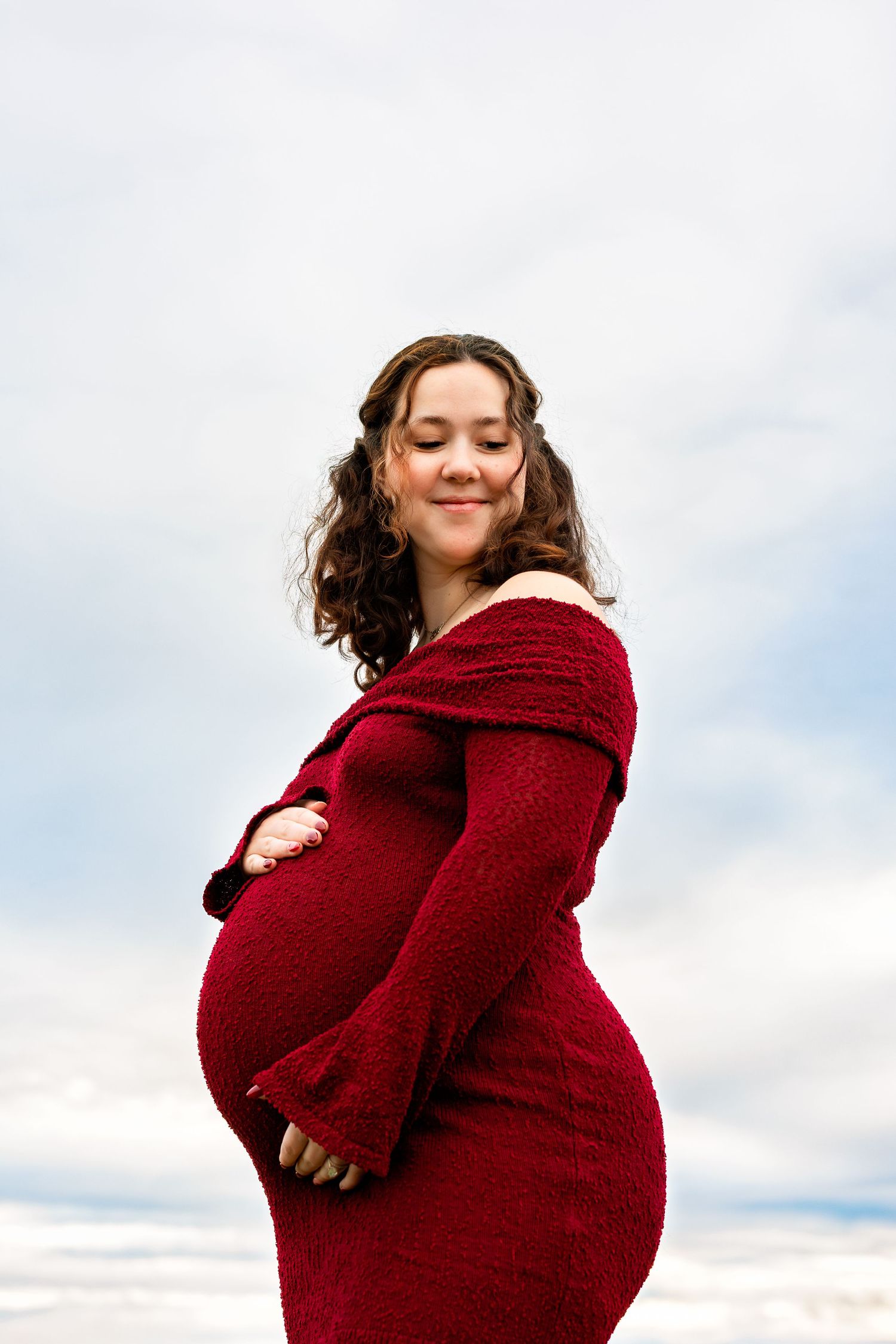 A pregnant woman in a burgundy sweater poses outdoors against a cloudy sky in a series of maternity photos.