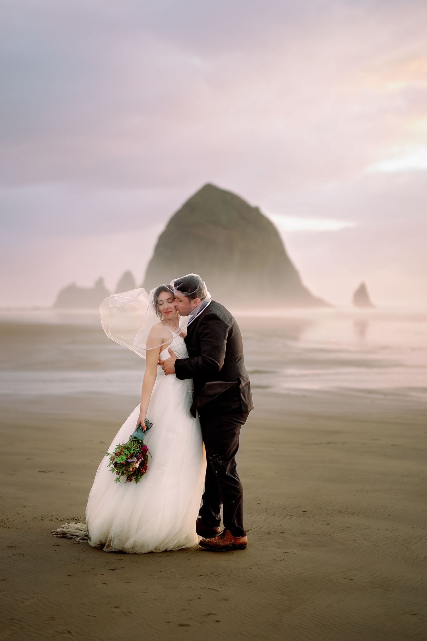 A couple shares an intimate moment on a misty beach with Haystack Rock at Cannon Beach, Oregon in the background.