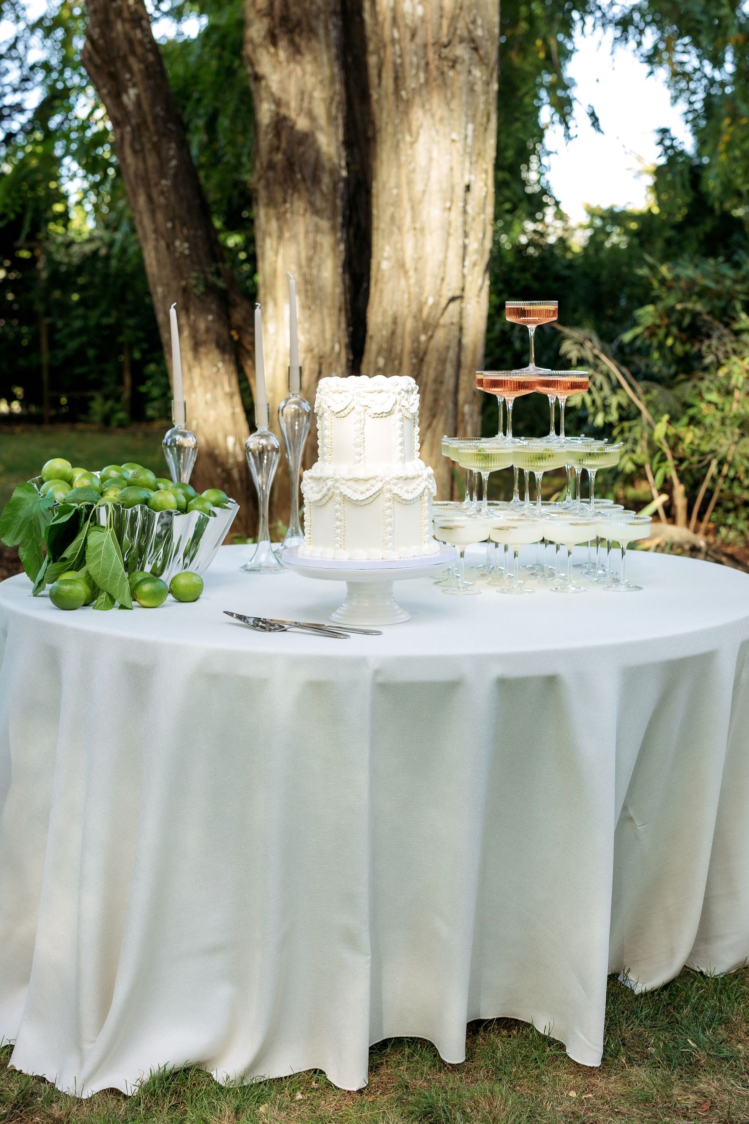 Elegant outdoor wedding reception setup with white tablecloths and glassware arranged under tall trees.