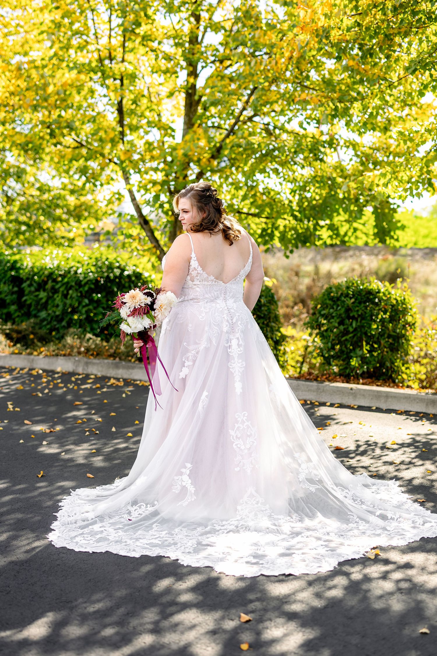 A bride in a lace wedding gown with a long train holds a bouquet against a backdrop of autumn trees.