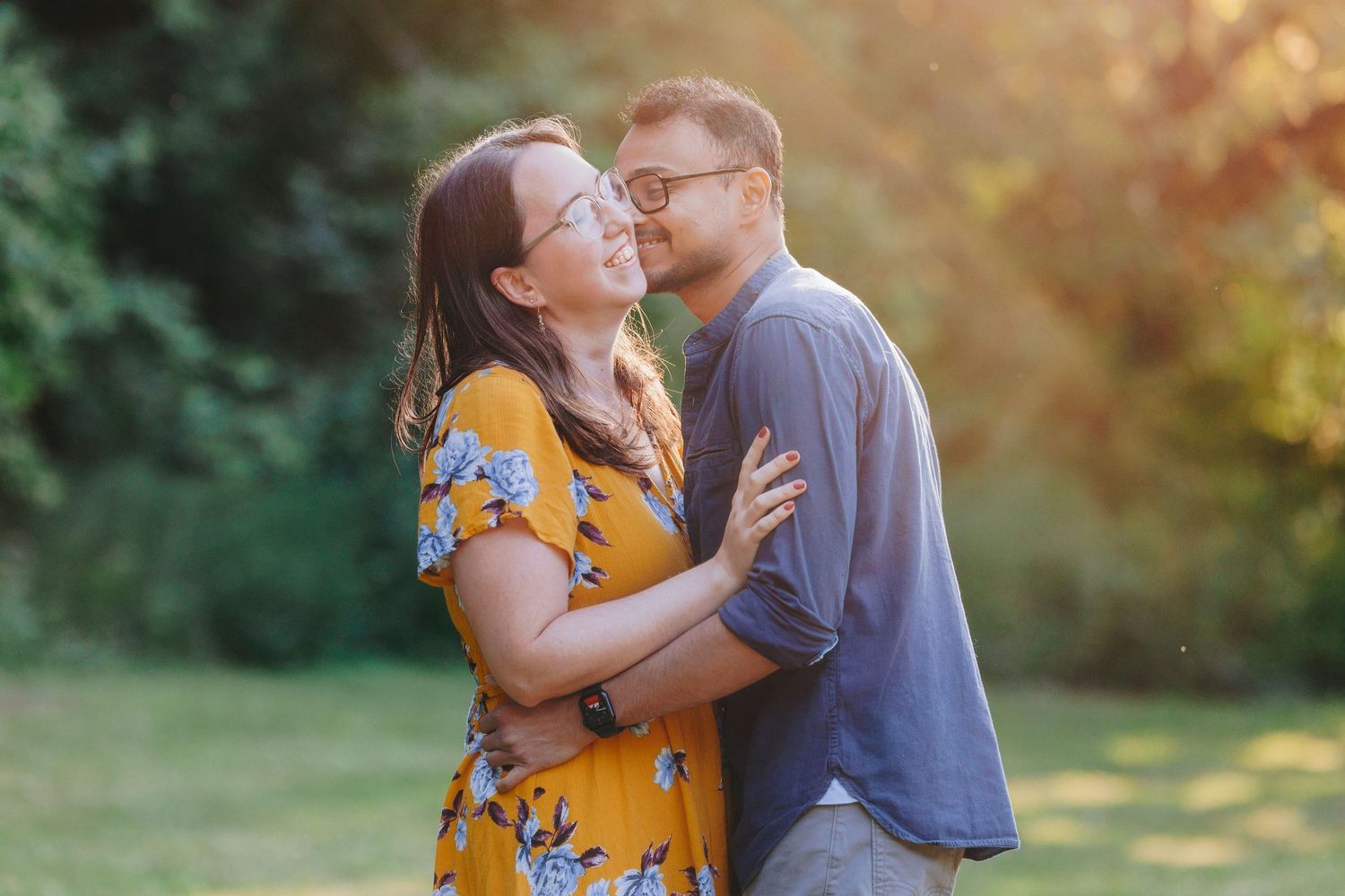 A couple embraces outdoors in warm sunset light, with her wearing a yellow floral dress and him in a blue shirt.