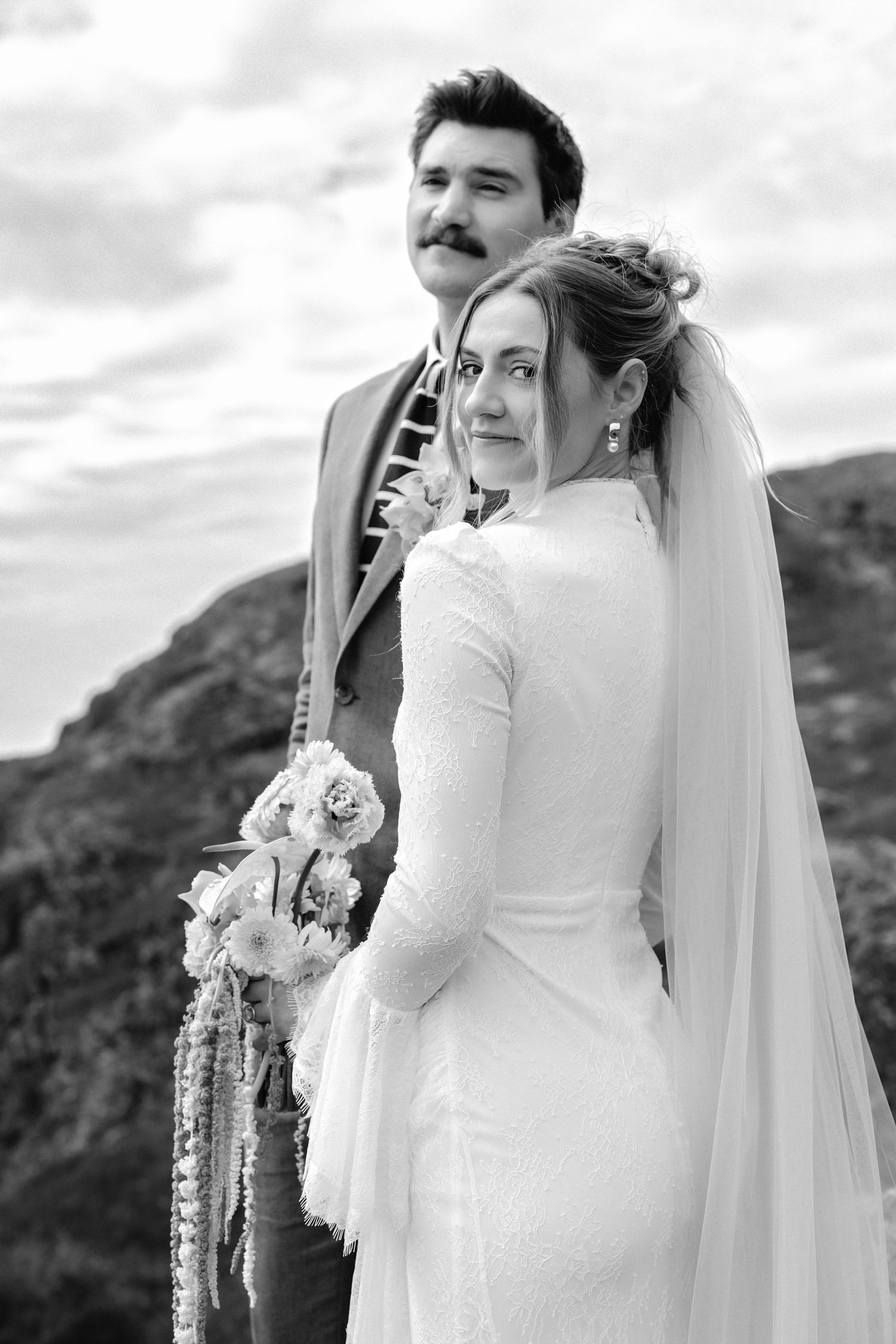Black and white photo of a bride holding a floral bouquet and groom with mustache posing outdoors with rocky hills behind.