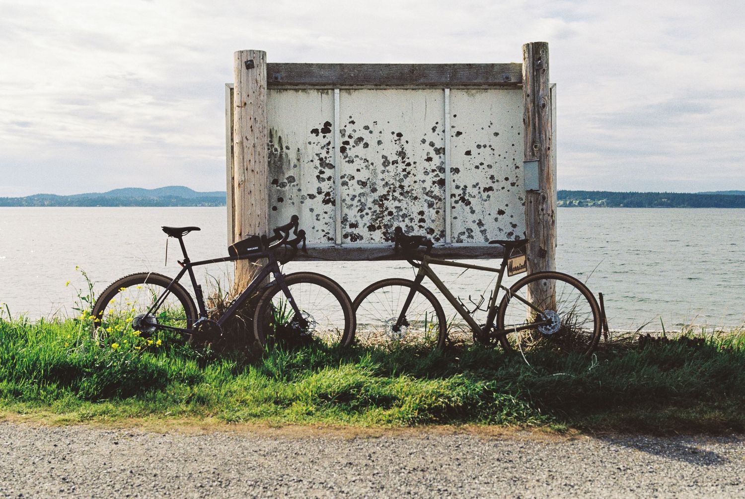 Two rusty vintage bicycles leaning against a weathered white structure beside a calm waterway.