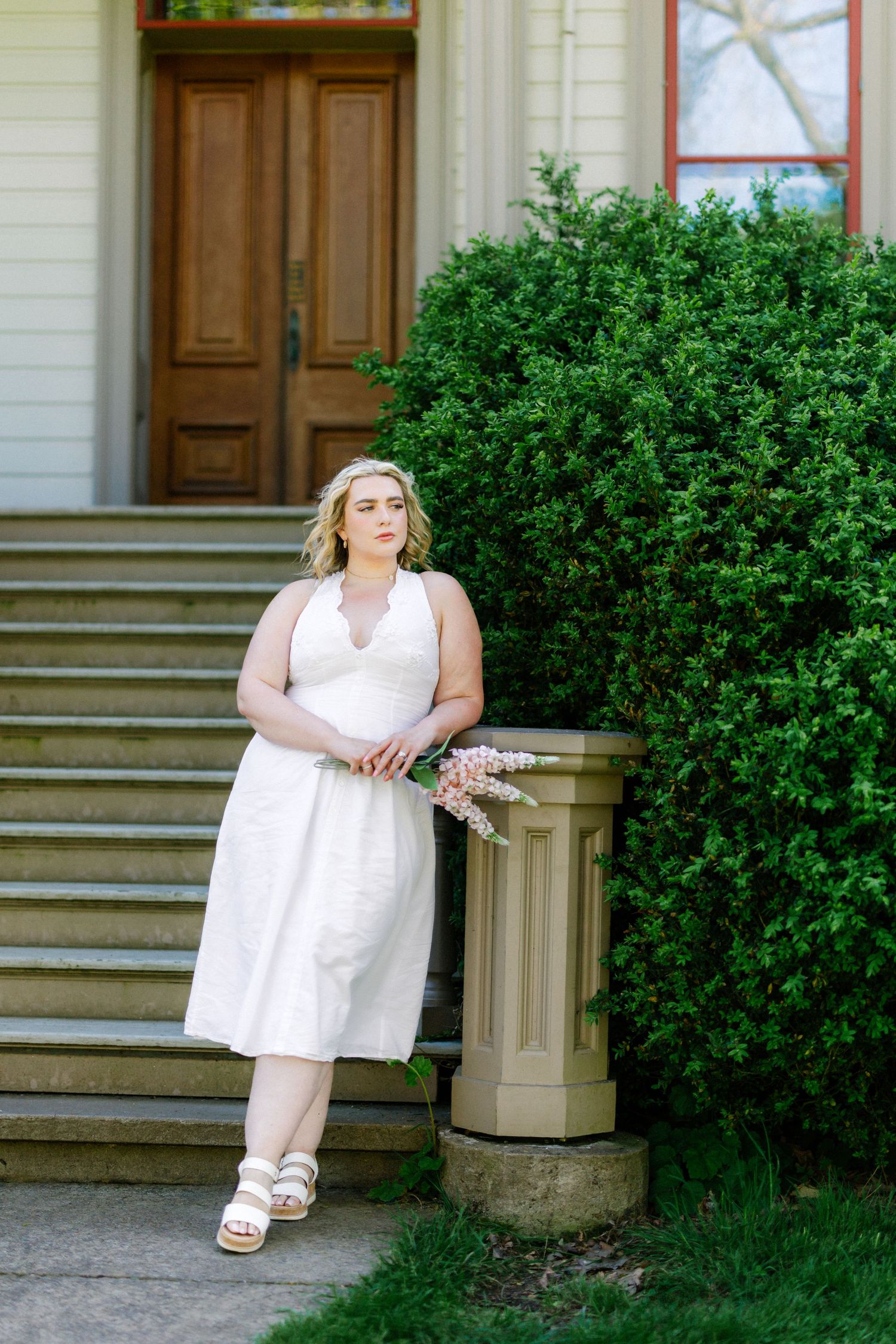 Woman in white dress holding flowers poses by stone pillar and green bushes outside historic building.
