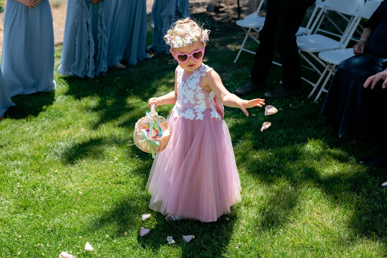 Adorable flower girl in pink tulle dress and sunglasses tossing petals at an outdoor wedding ceremony.