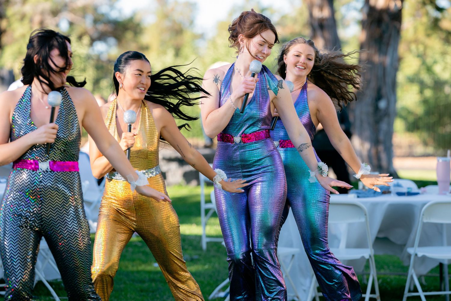 Four women in colorful metallic outfits dance energetically at outdoor wedding reception.