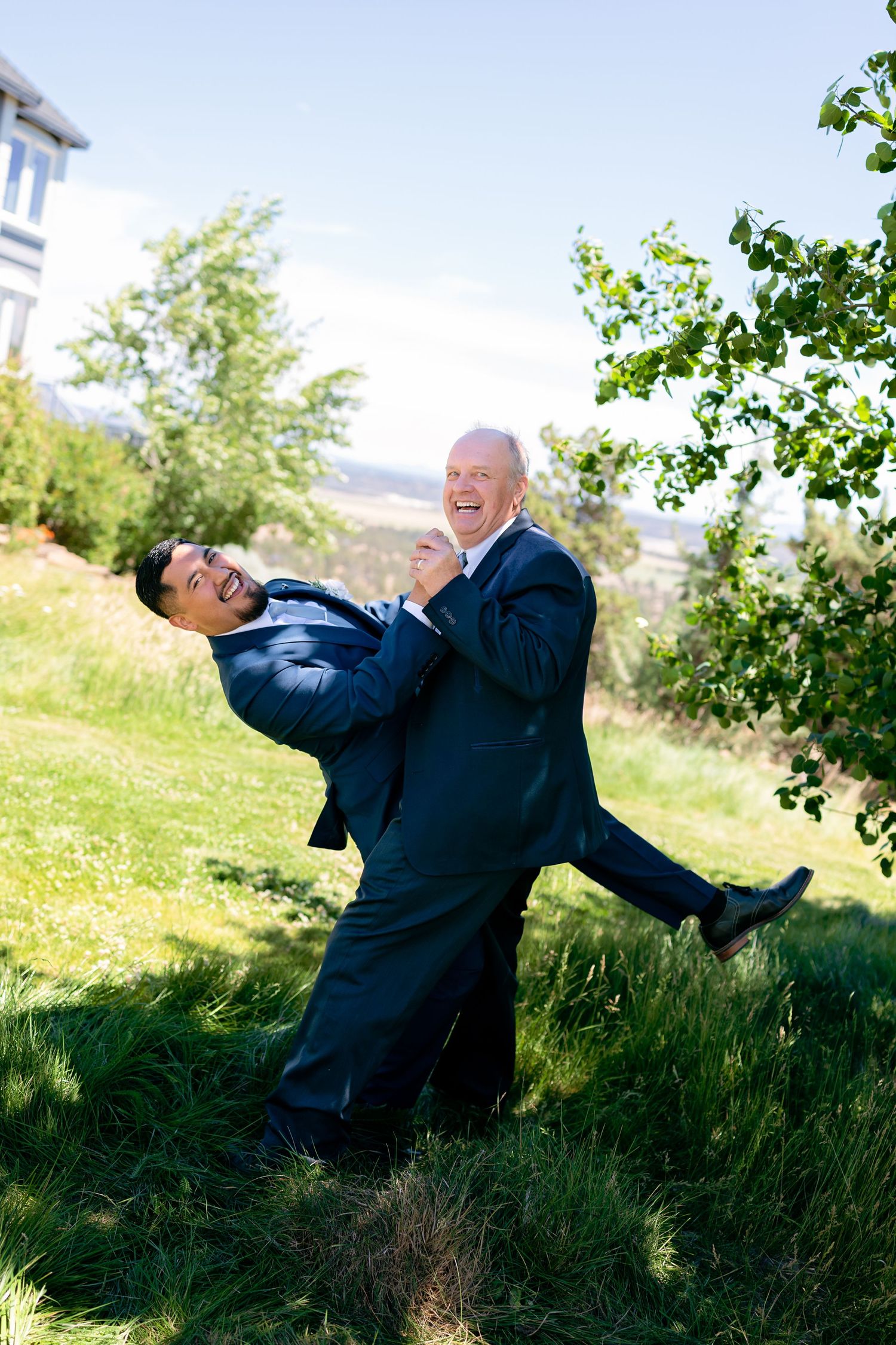 Two men in dark suits laugh joyfully as one lifts the other in a sunny outdoor setting.