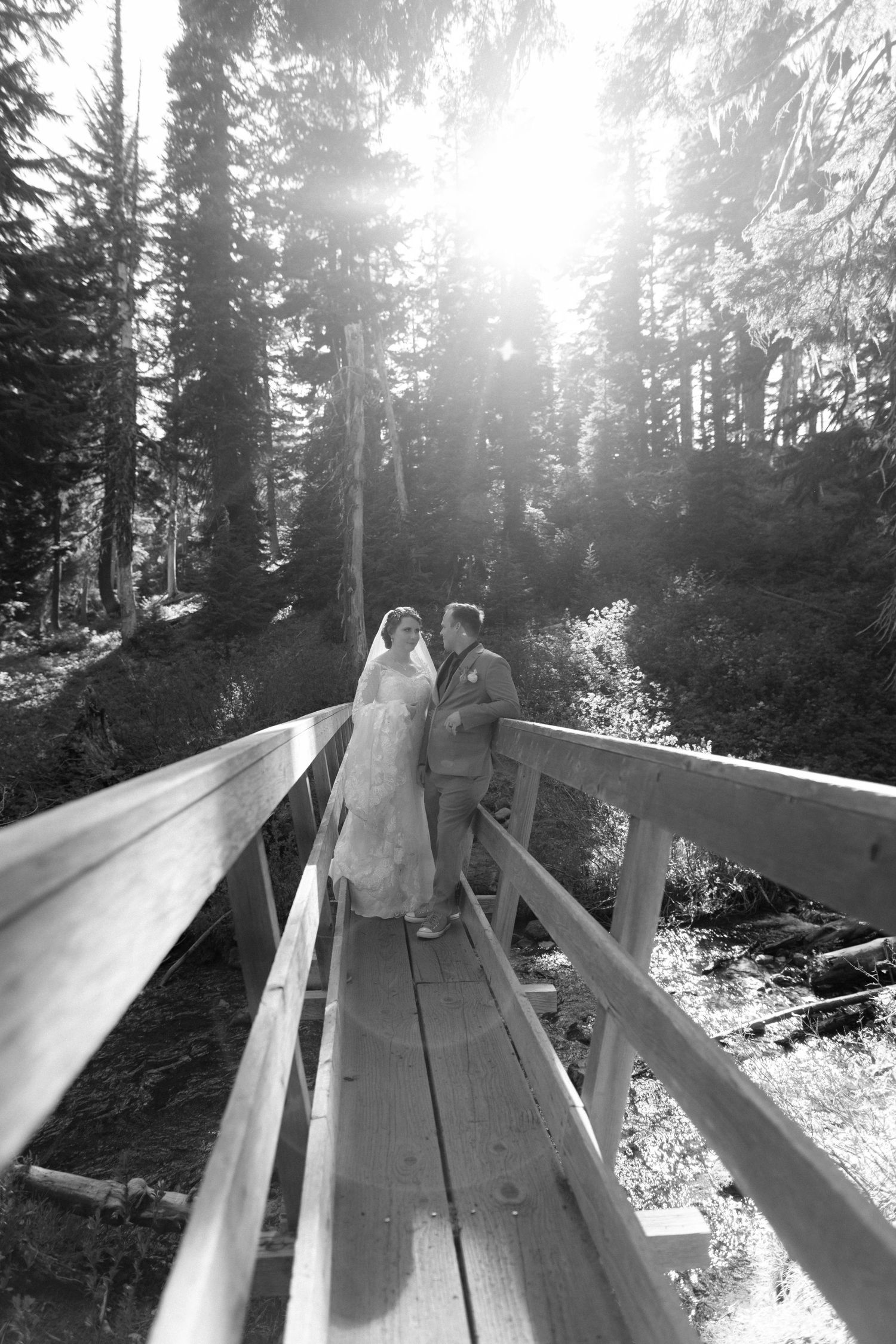 Black and white photo of bride and groom sharing a romantic moment on a wooden bridge in a forest.