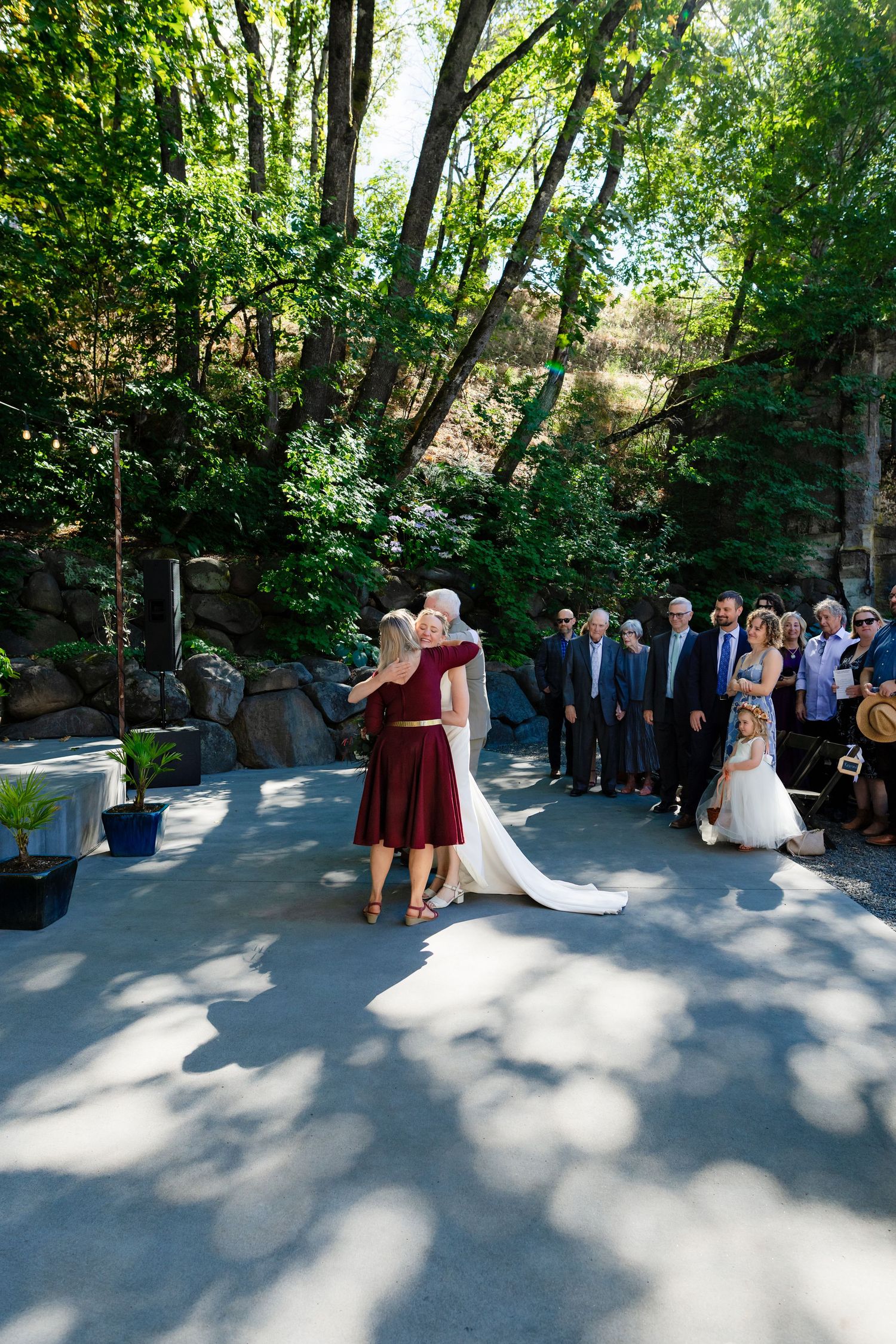 Bride embraces woman in burgundy dress during outdoor wedding ceremony surrounded by lush green trees.