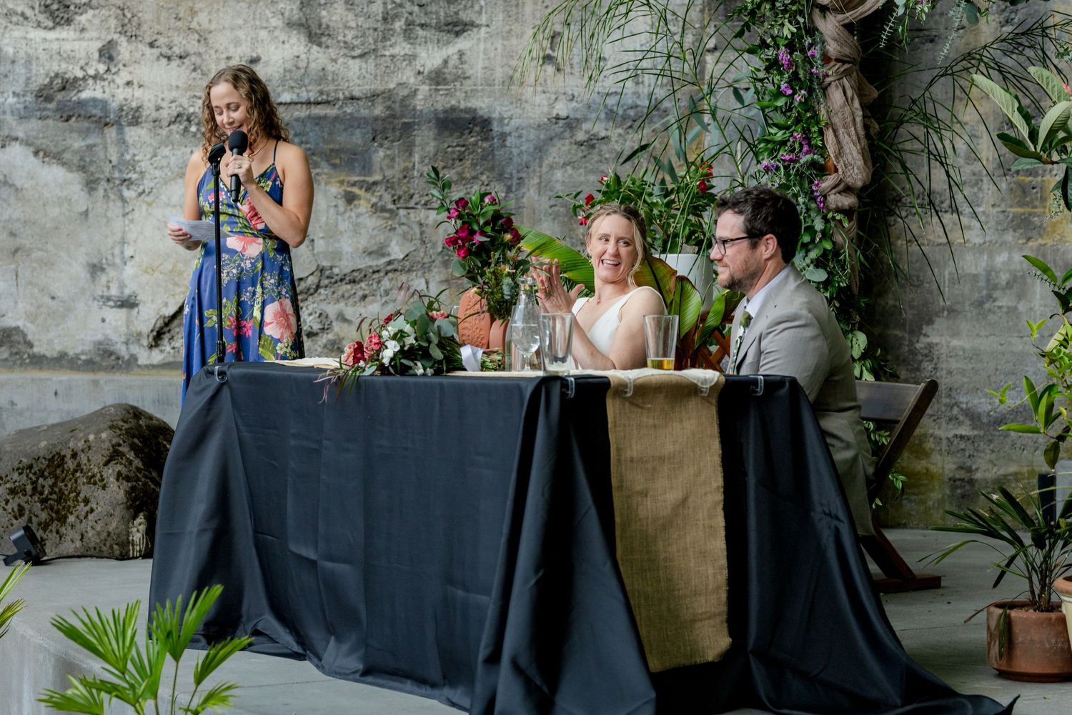 Wedding guest gives toast as bride and groom sit at floral decorated sweetheart table.
