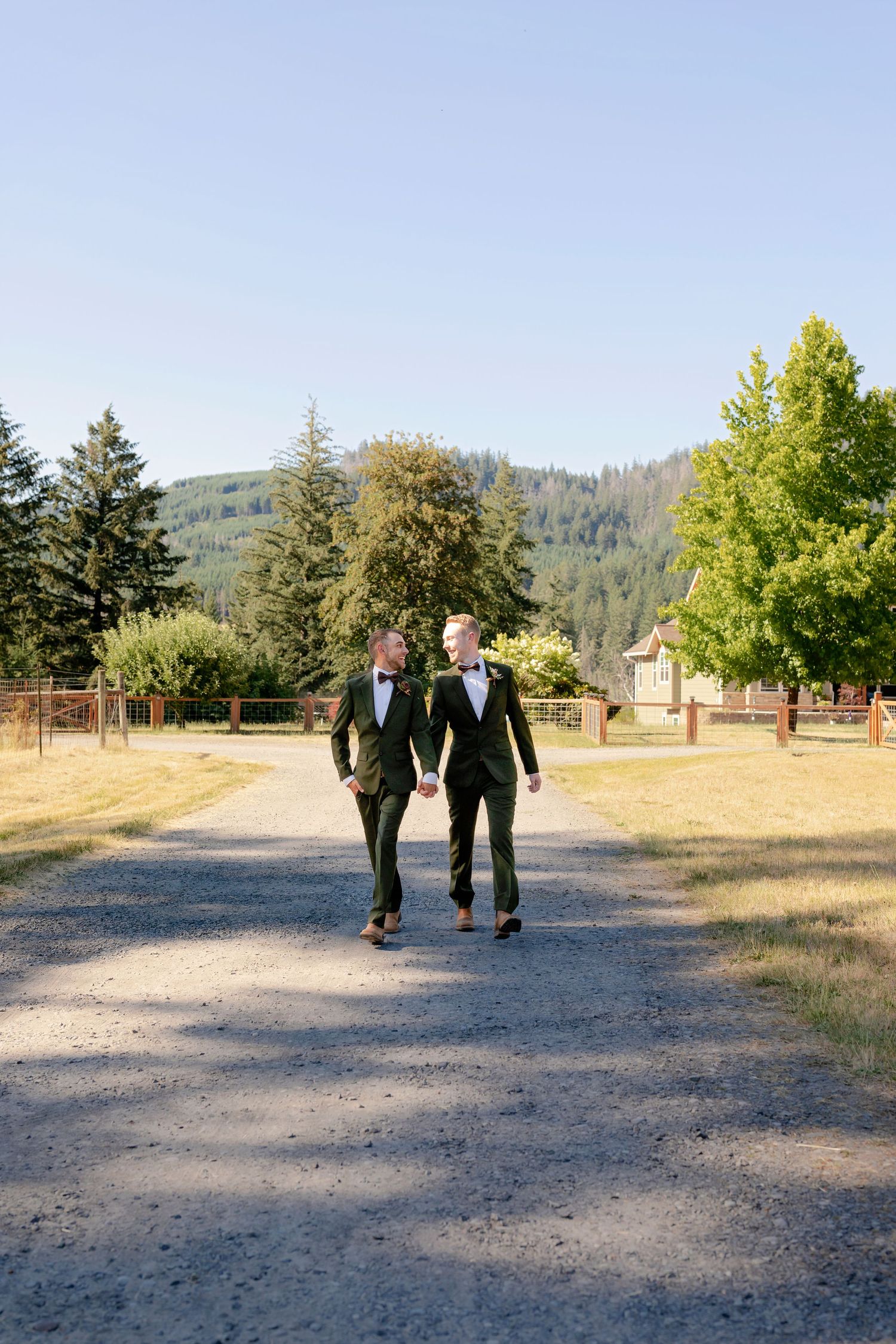 Two grooms in matching dark suits and bow ties walk hand in hand on a scenic country path.