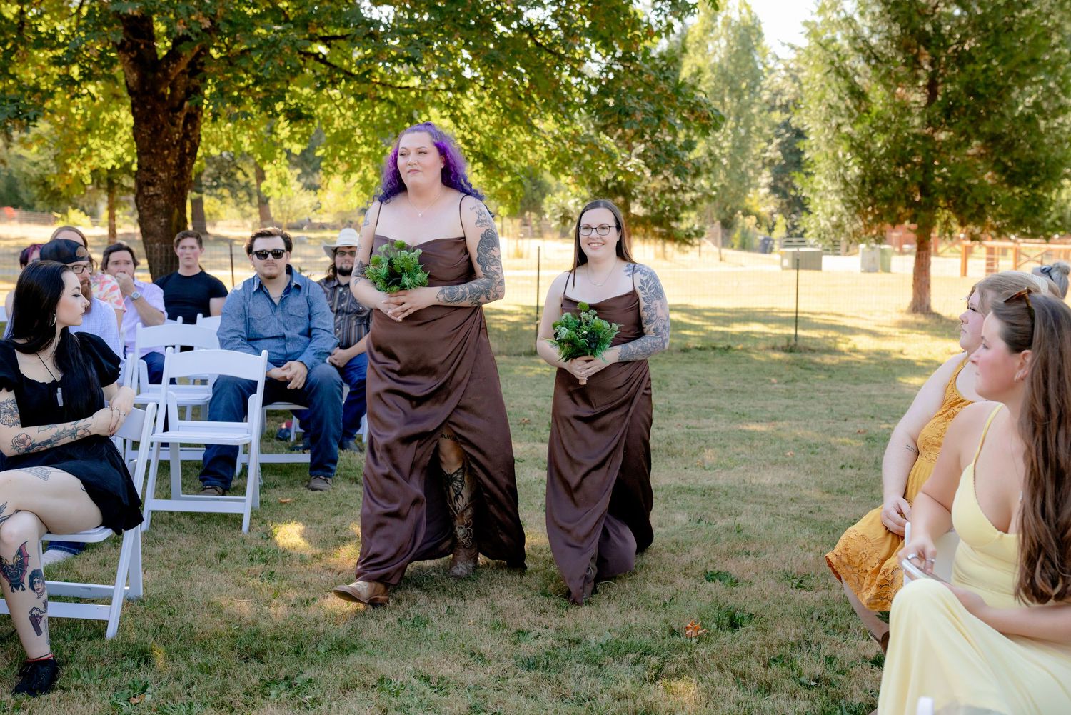 Two bridesmaids in brown dresses carrying green bouquets walking down outdoor wedding ceremony aisle.