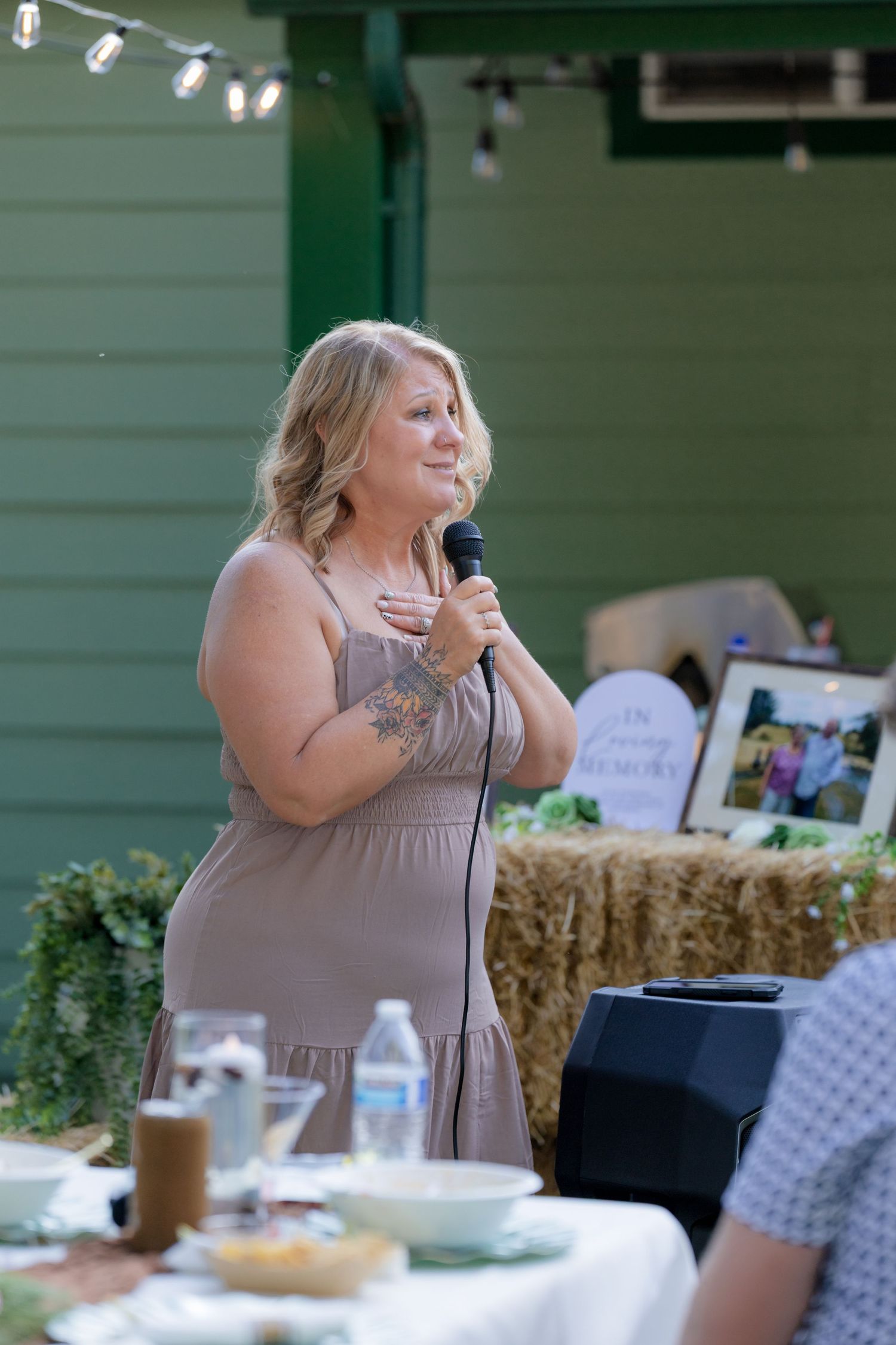 Blonde woman in taupe dress giving a heartfelt speech with microphone at outdoor wedding reception.