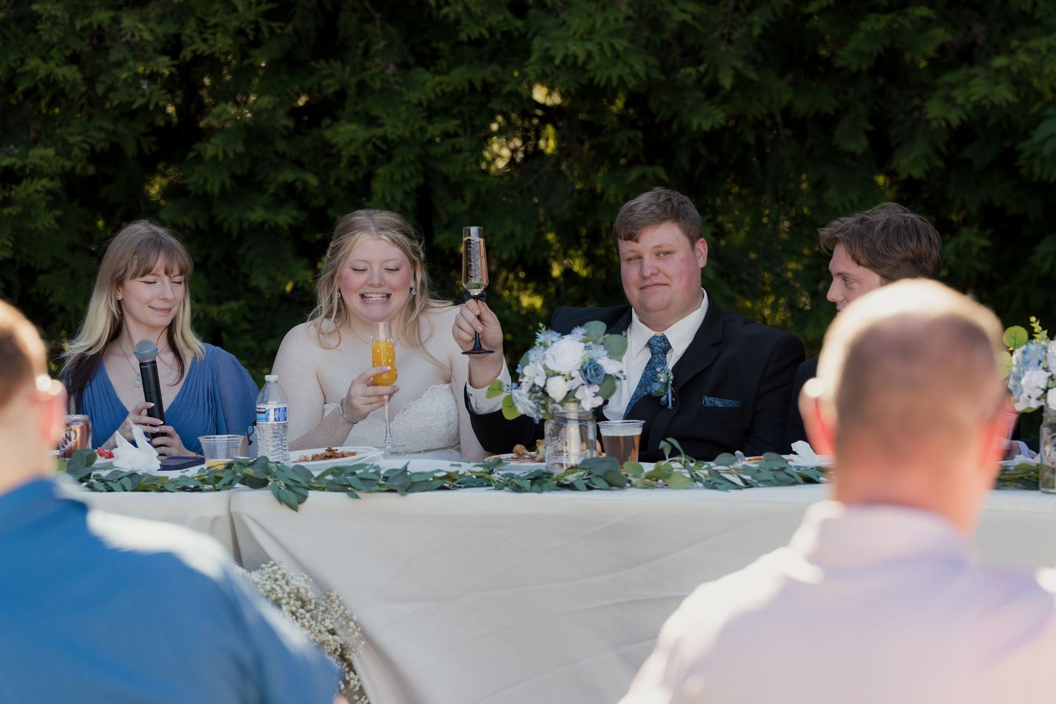 Bride and groom raising champagne glasses during outdoor wedding reception toast at head table.