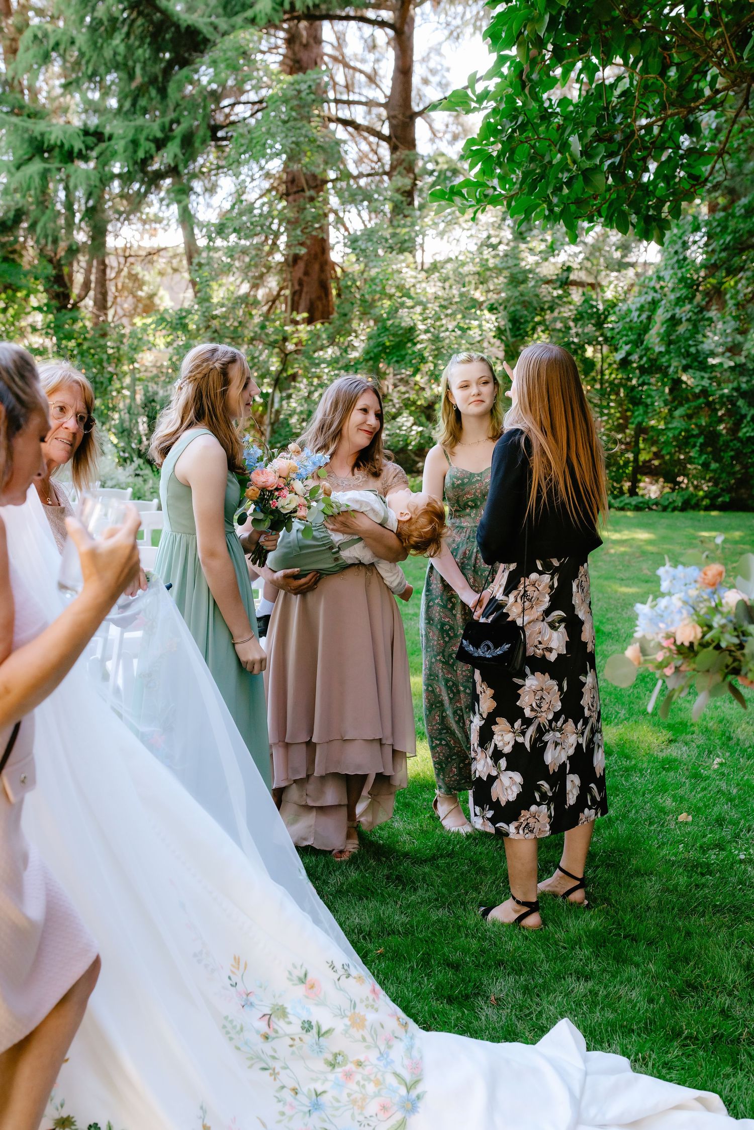 Bridesmaids in sage and taupe dresses gather on green lawn holding bouquets beside bride's embroidered wedding gown train.