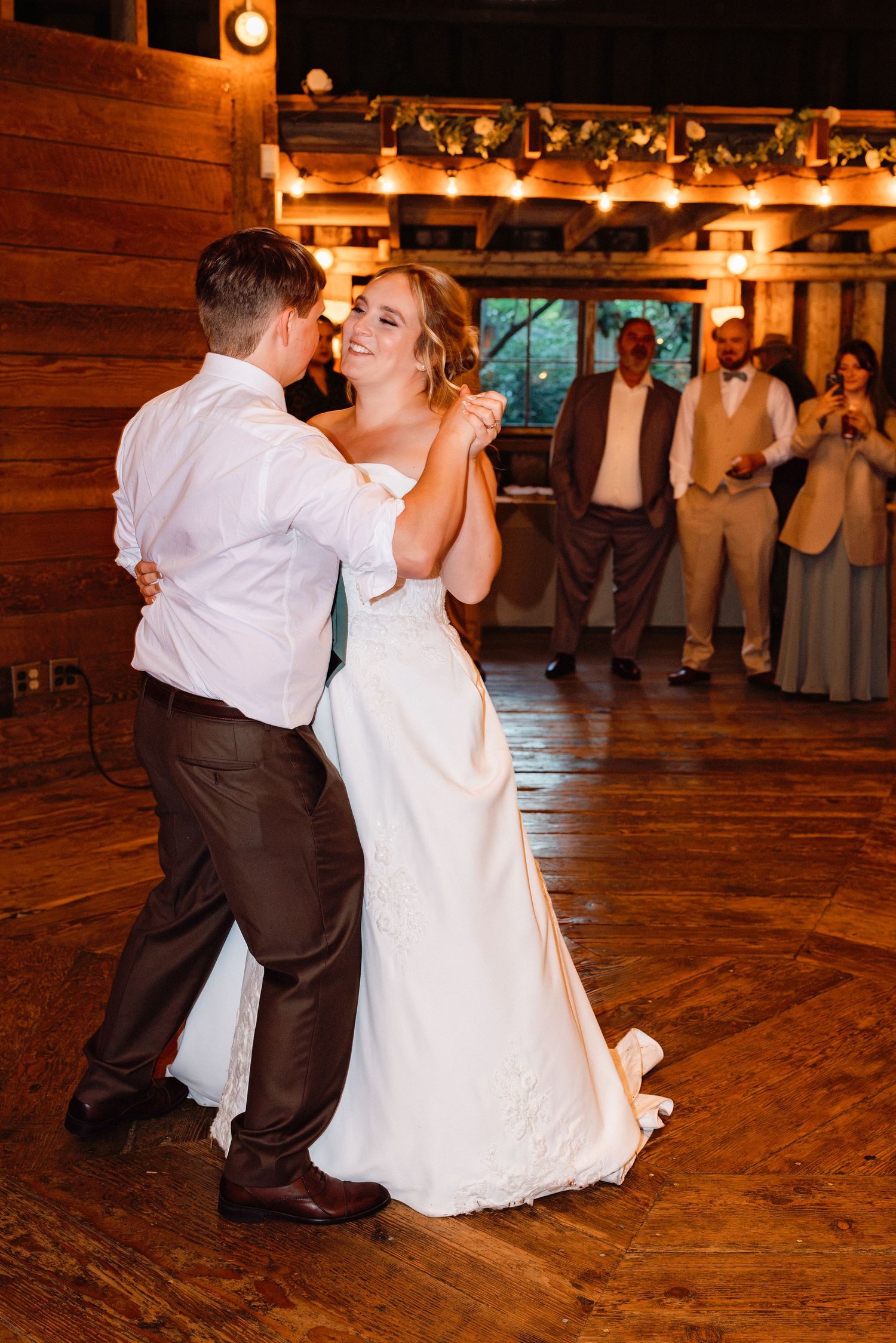 Smiling bride in white gown dances with groom on wooden dance floor in warmly lit barn.