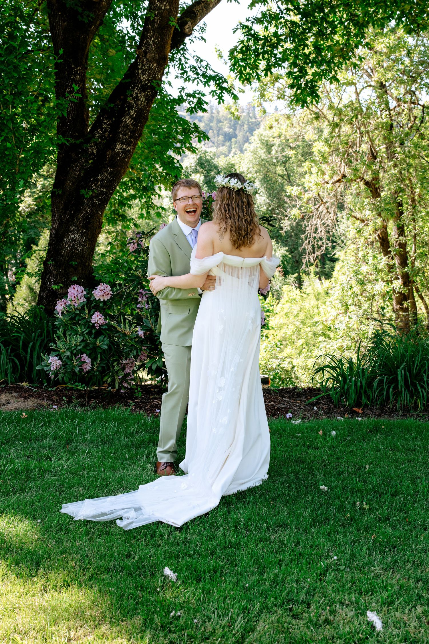 Groom in sage suit laughs joyfully during first look with bride in white gown and floral crown in garden.