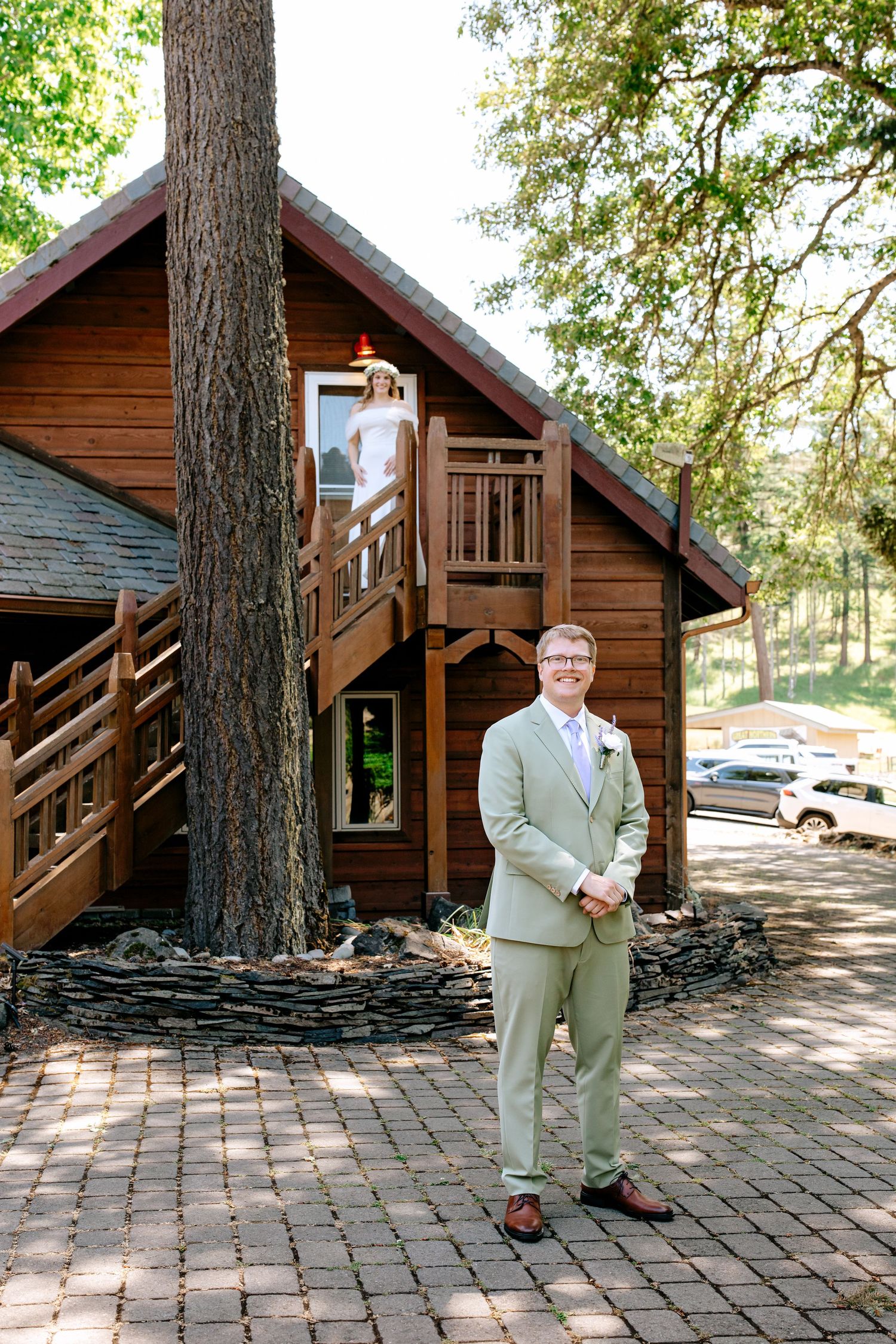 Groom in sage suit stands before rustic wooden cabin as bride watches from staircase above during first look.