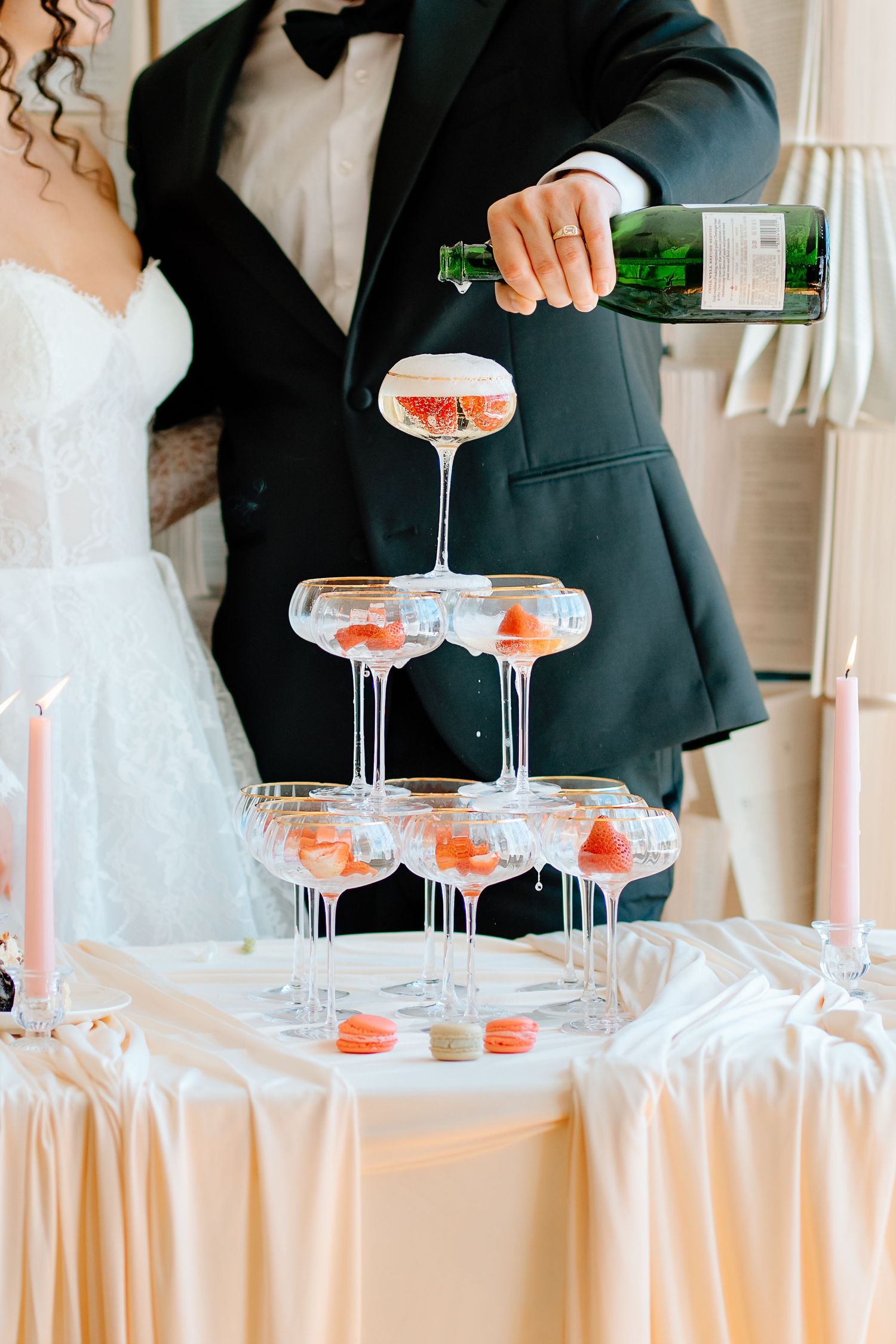 Bride and groom pouring champagne into a elegant tiered glass tower with pink macarons at a wedding reception.
