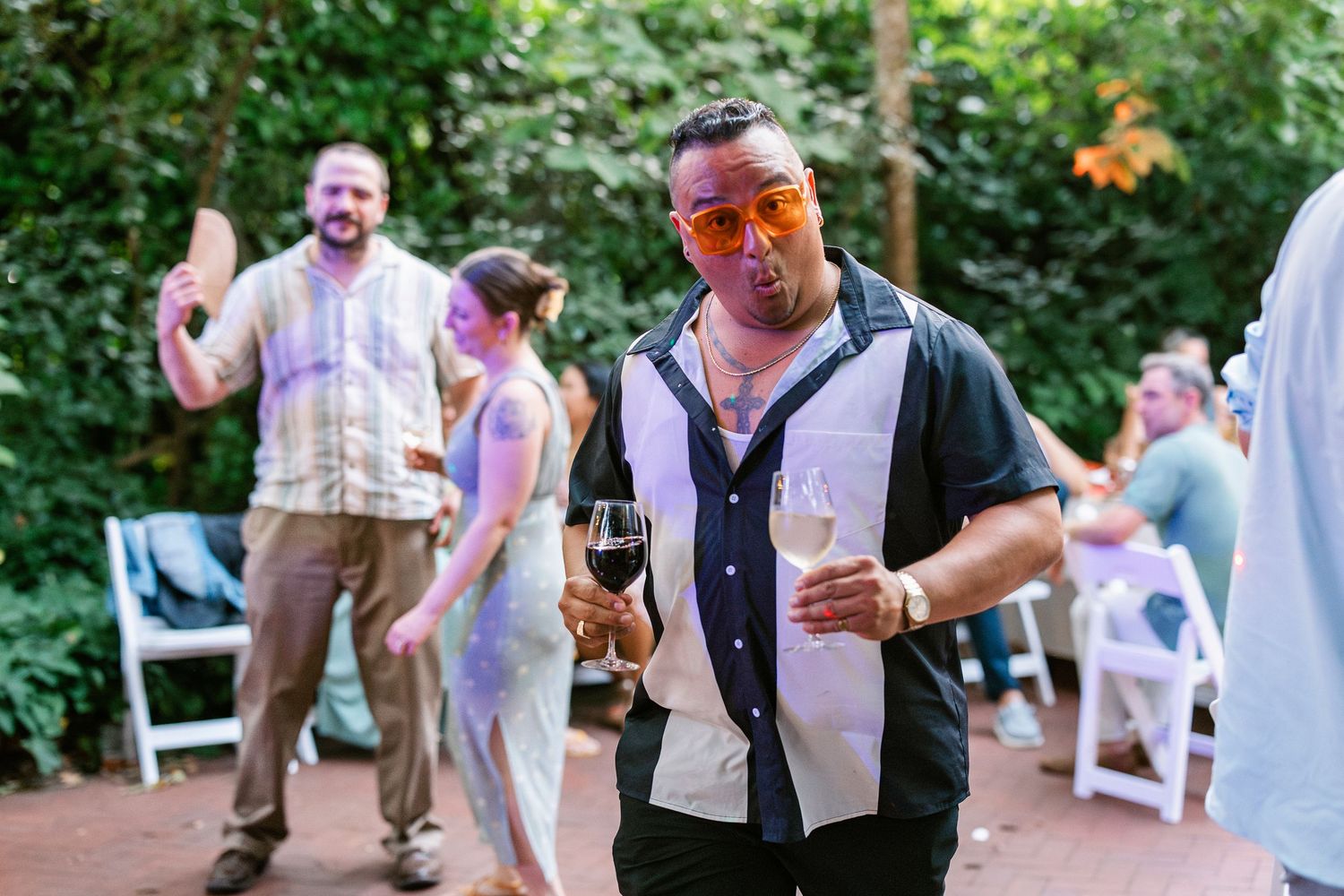 Man in orange sunglasses holding wine glass at outdoor garden party with guests socializing in background.