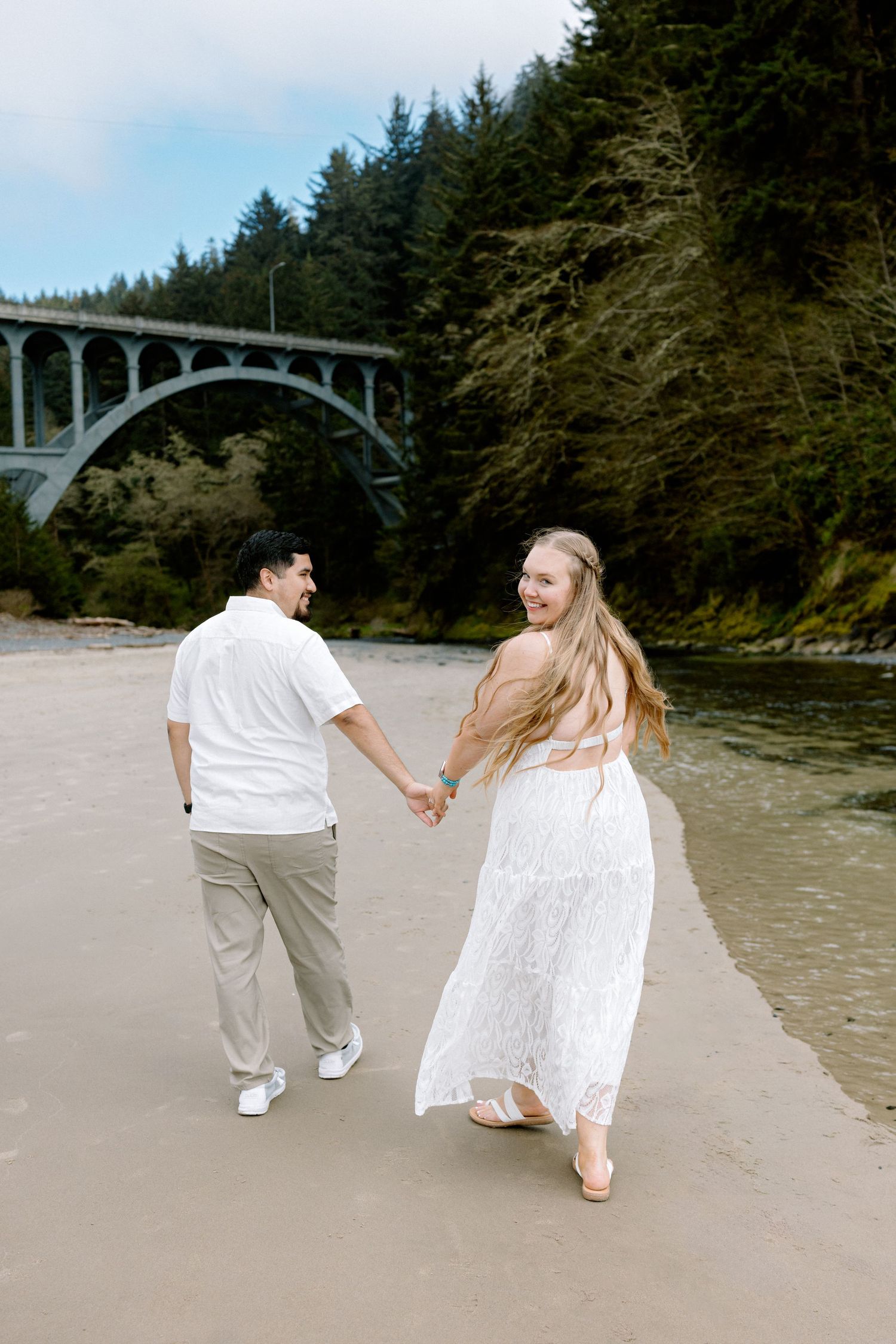 Couple holding hands on sandy beach in white outfits, smiling near a river with an arched bridge and forest backdrop.